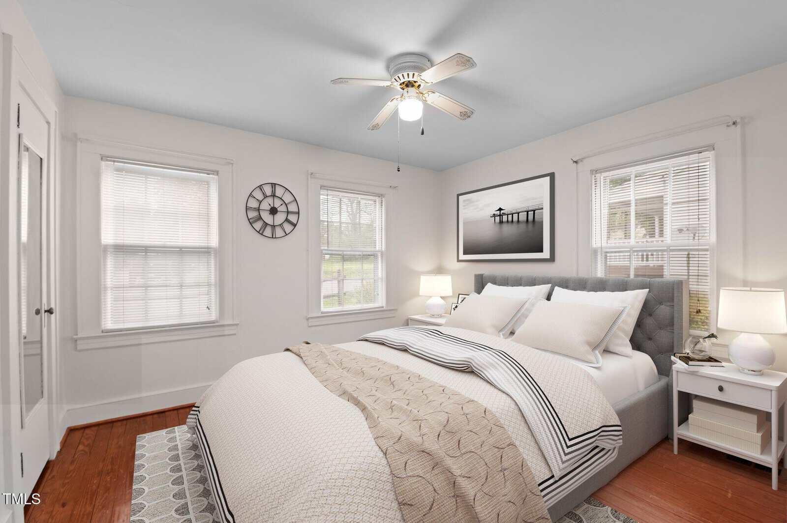 A cozy bedroom with white walls and hardwood floors features a bed with white and beige bedding. There are two windows with blinds, a ceiling fan, a clock on the wall, a landscape picture, and two bedside tables with lamps.