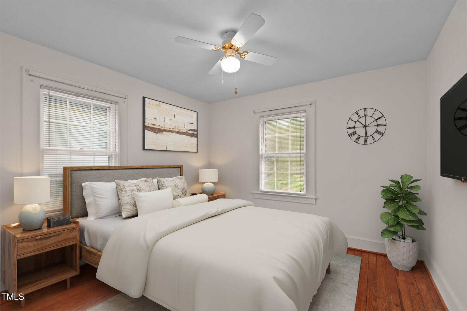 A bedroom with a bed featuring white bedding and wooden headboard. Two wooden nightstands with lamps are on each side. A ceiling fan is above, with a wall-mounted TV, a potted plant, and a decorative clock on the walls. Natural light enters through two windows.