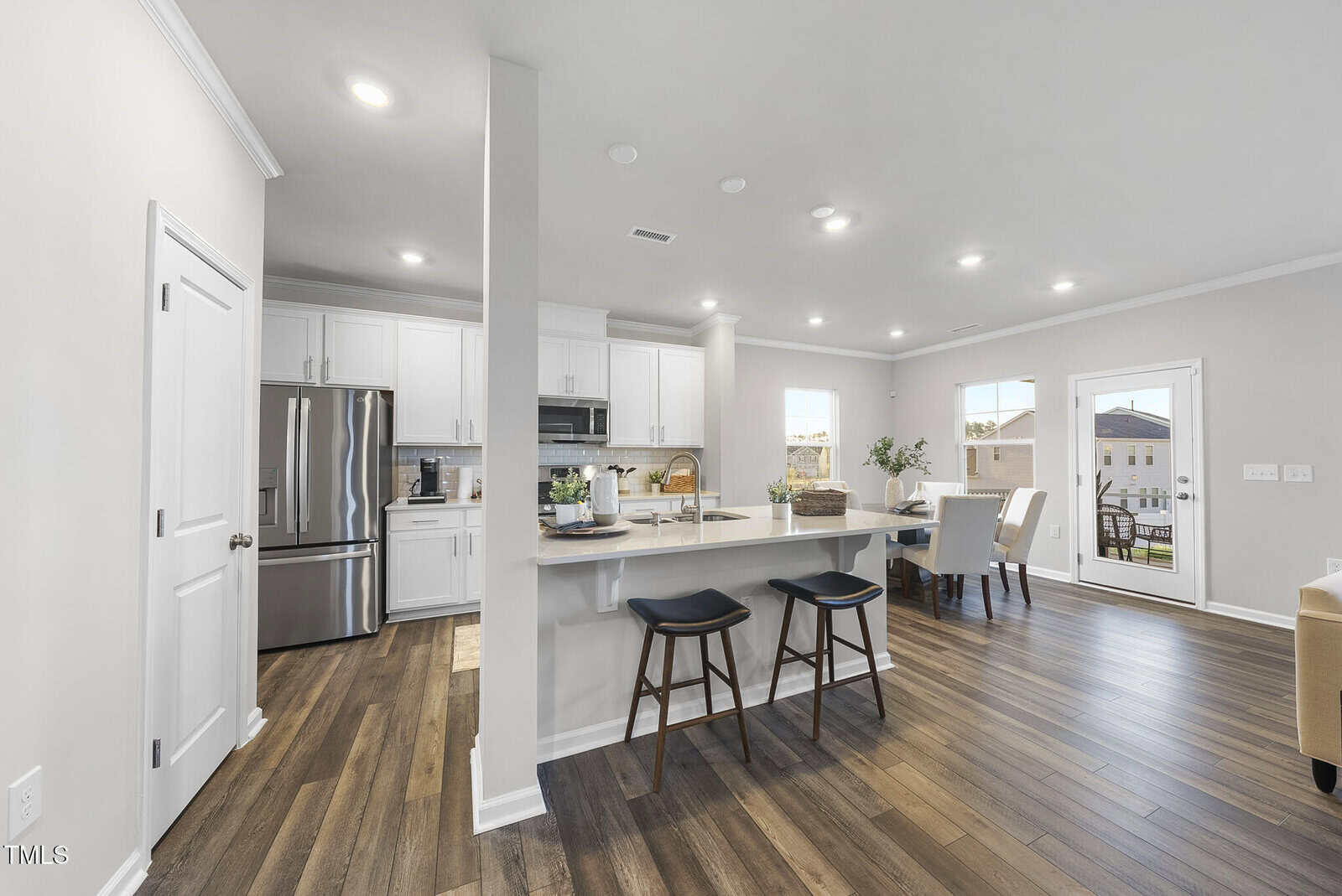 Modern kitchen with white cabinets, stainless steel appliances, an island with two black stools, and wood flooring. The space opens to a dining area and living room with large windows and a glass door to the outside.
