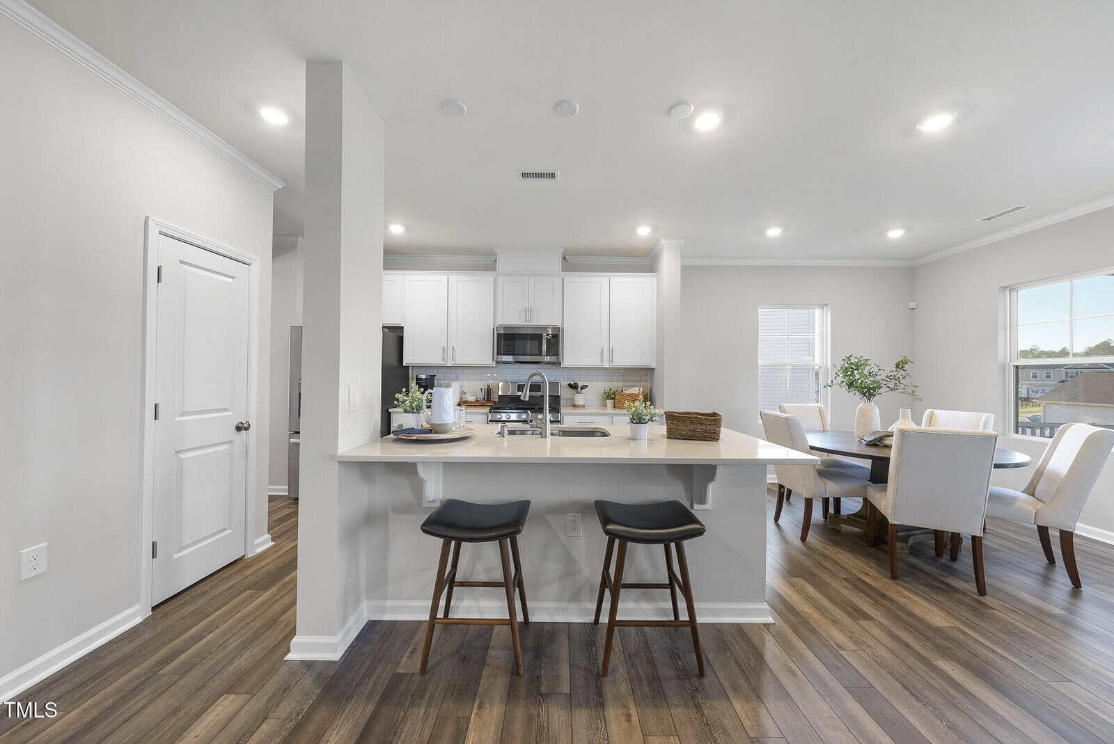 Modern kitchen with white cabinets, stainless steel appliances, and a breakfast bar with two black stools. Adjacent dining area features a wooden table and upholstered chairs. Wood flooring throughout the space.