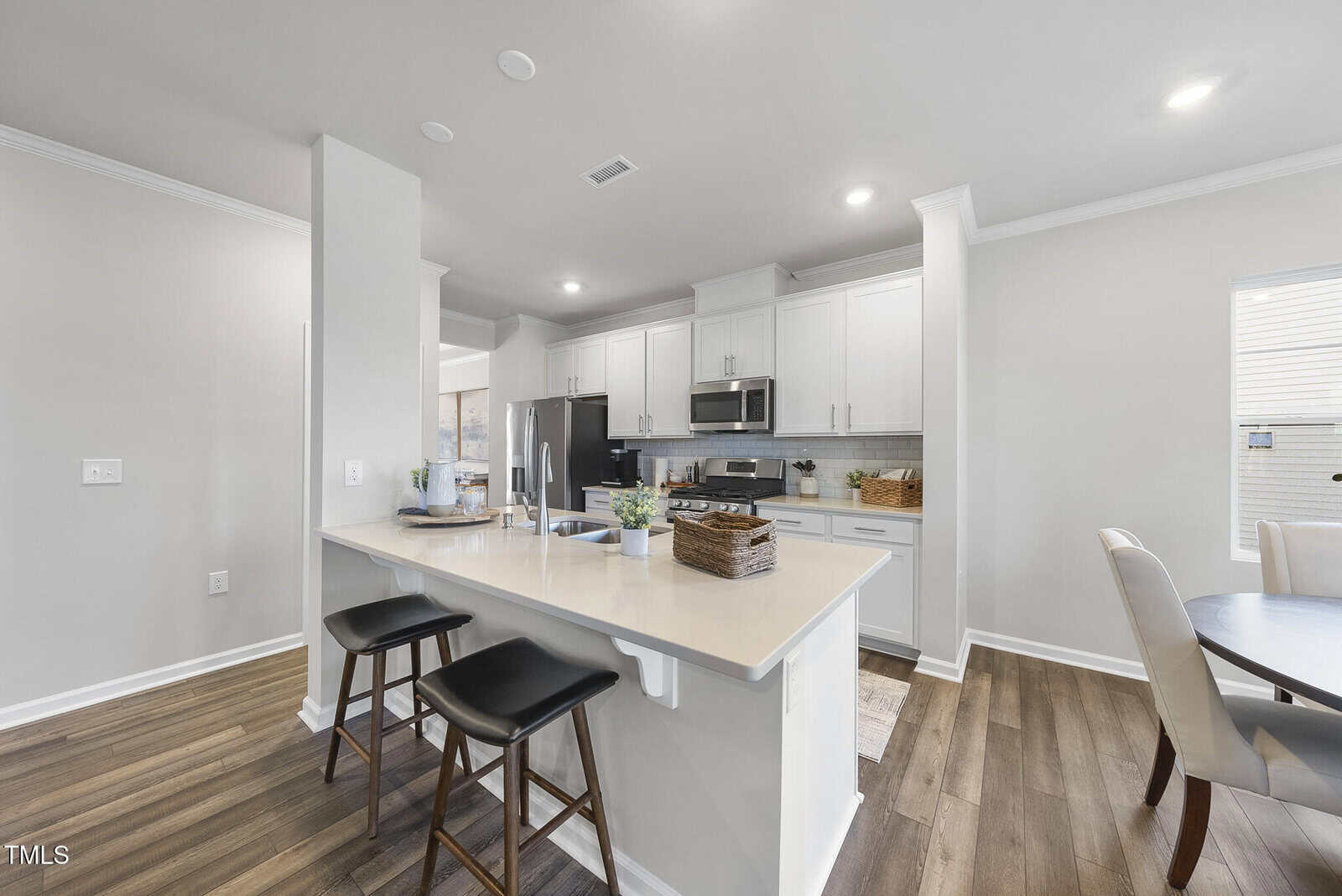 A modern kitchen with white cabinets, stainless steel appliances, a large island with bar stools, and a dining table. The room has light-colored walls and wood flooring, with natural light coming in from the window.