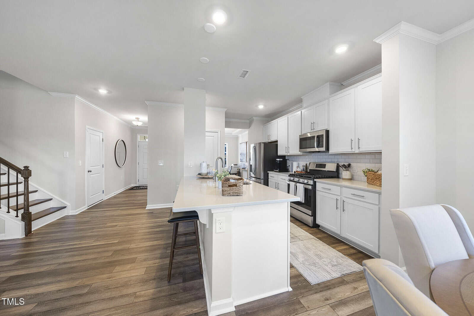 Modern kitchen with white cabinets, stainless steel appliances, and a light-colored island with two black barstools. Wood flooring, neutral walls, and stairs are visible in the background. The space looks clean and organized.