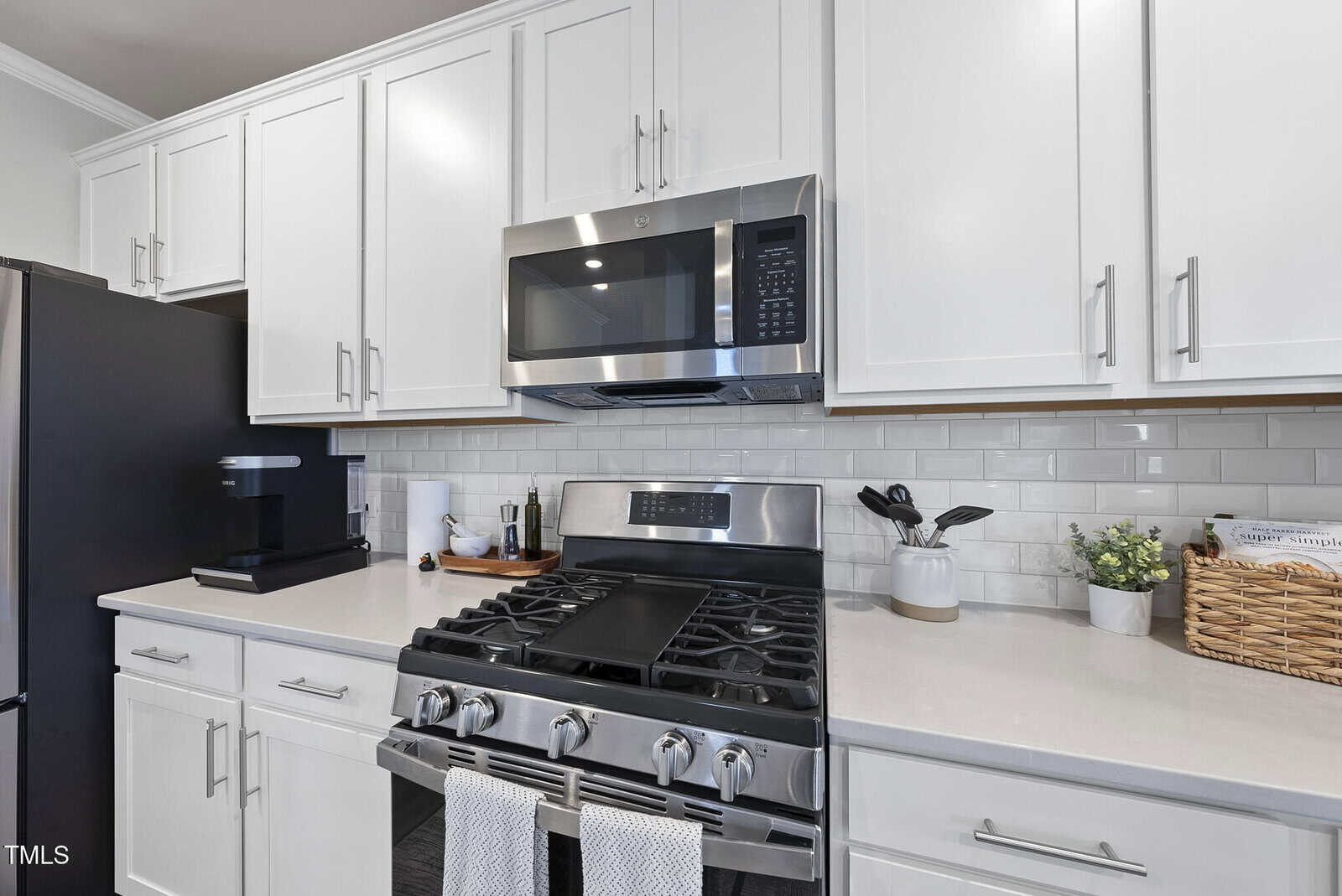 Modern kitchen with white cabinets, stainless steel gas stove and microwave, black coffee maker, utensils in a holder, a small plant, and a wicker basket on a light countertop with white subway tile backsplash.