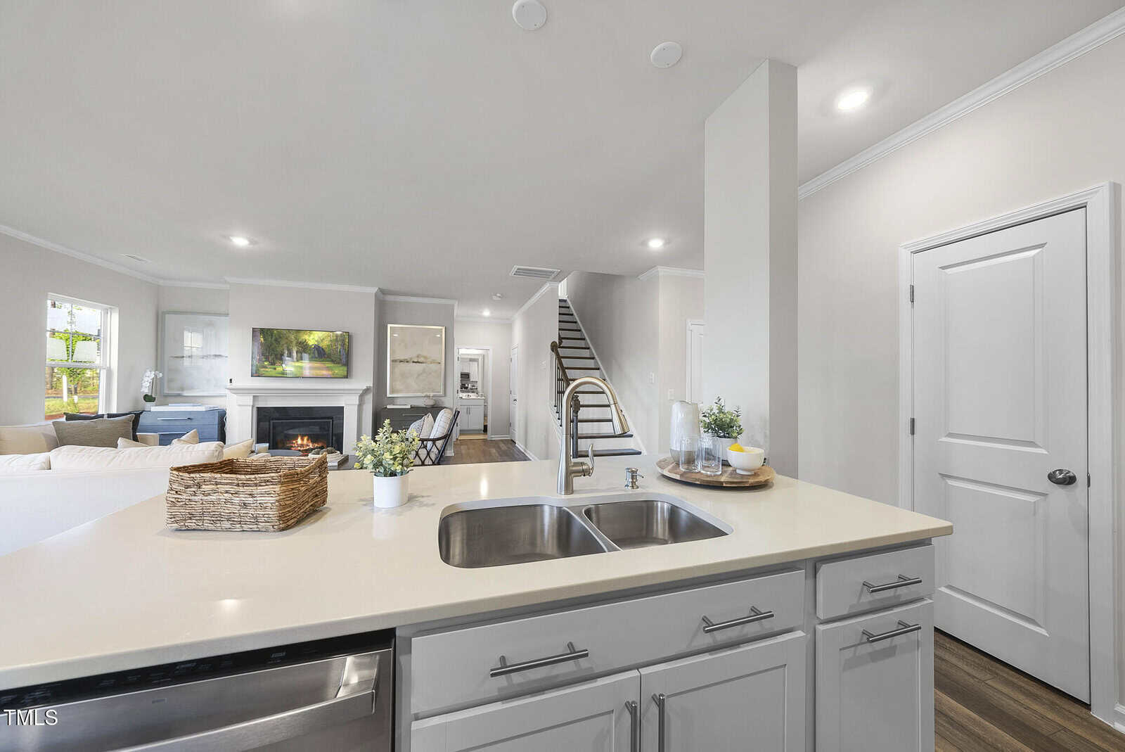 Modern kitchen with a double sink island, stainless steel appliances, and neutral decor. Living area with a fireplace and TV visible in the background. Staircase leads to upstairs. Wood floors and white walls throughout.