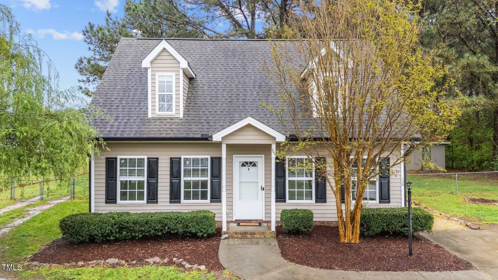 A two-story beige house with white trim and black shutters, featuring a front porch with a white door, surrounded by green shrubs and a tree, with a concrete walkway and a grassy yard.