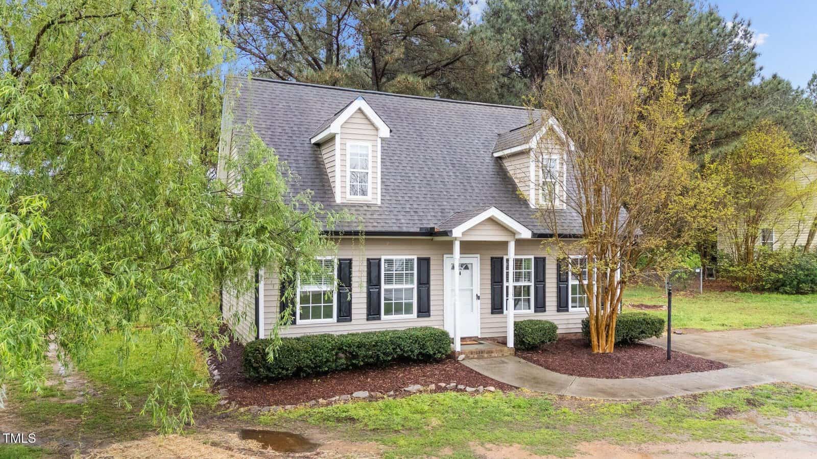 A two-story house with light siding, black shutters, and a dark shingle roof. It has dormer windows, a covered front porch, shrubs, and a curved concrete path leading to the front door, surrounded by trees and grass.