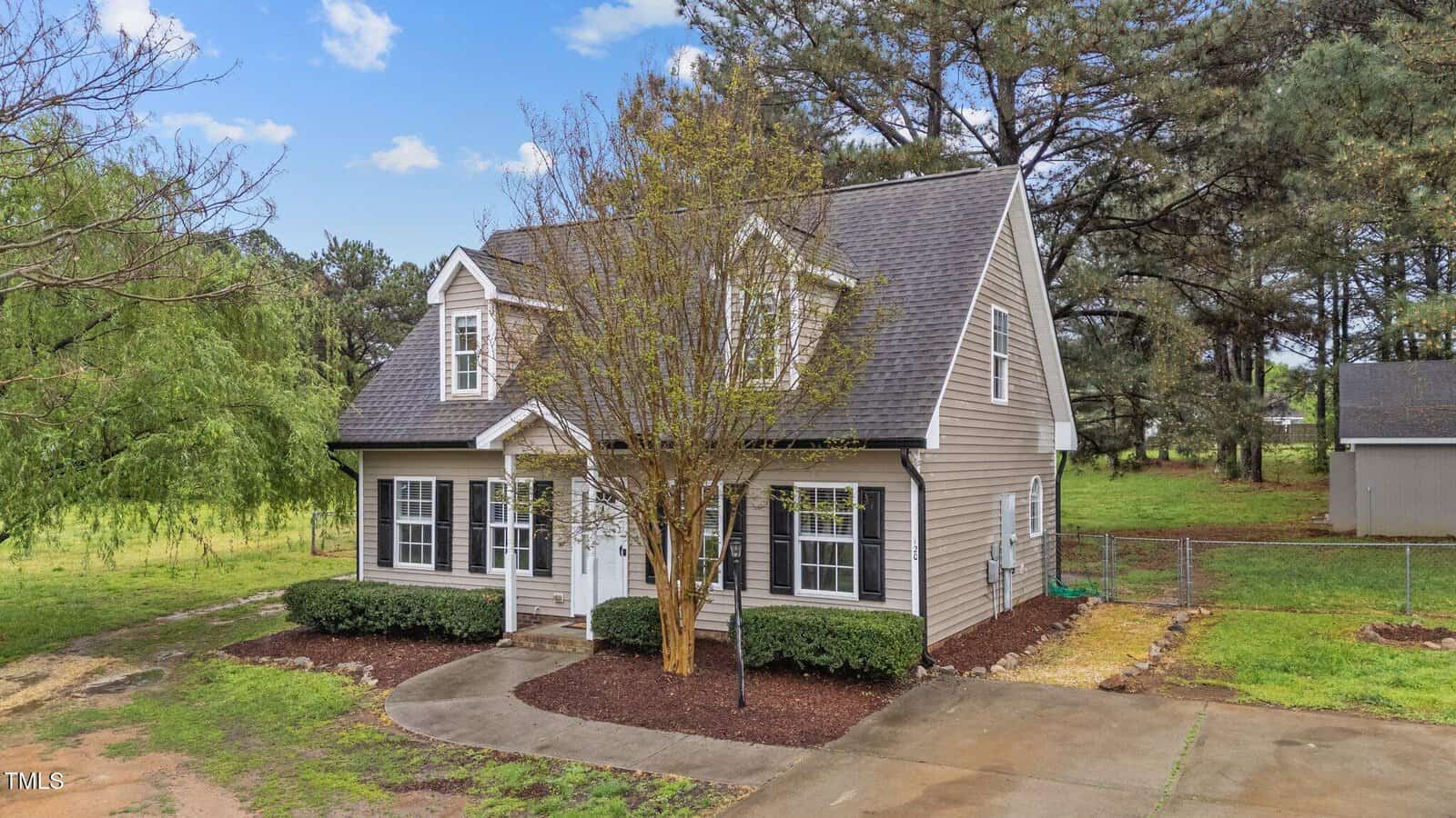 A two-story beige house with white trim and a dark gray roof sits on a grassy lot surrounded by trees. There is a small tree in front, hedges along the front wall, and a circular driveway with a concrete path to the door.