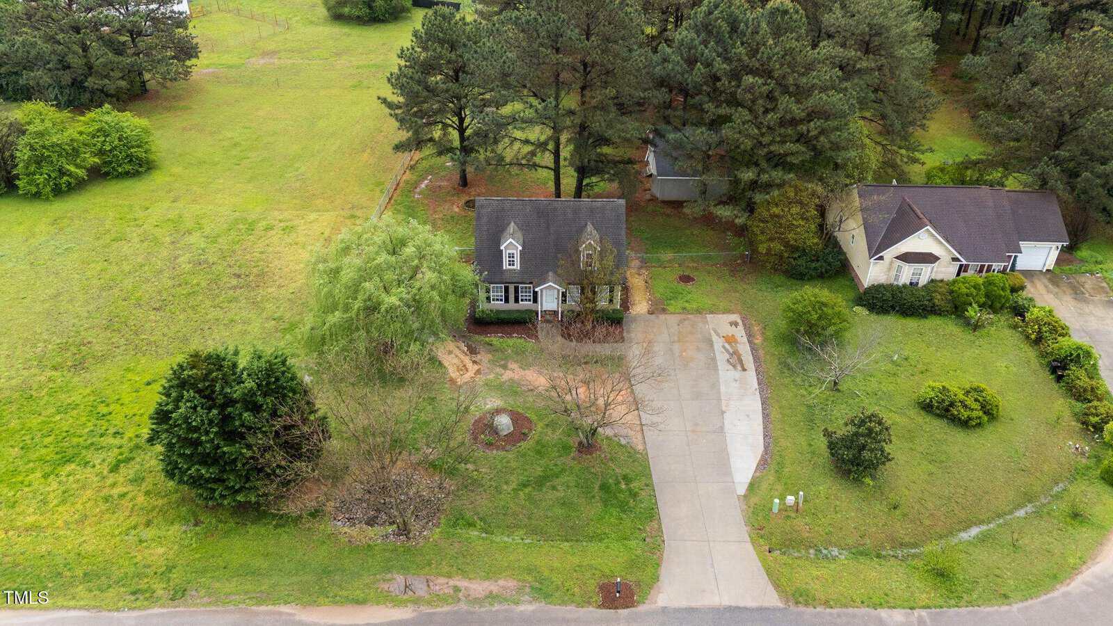Aerial view of a suburban home with a grey roof, surrounded by trees and grassy lawns. A concrete driveway leads to the house, and a neighboring house is visible to the right. The background includes open green space.