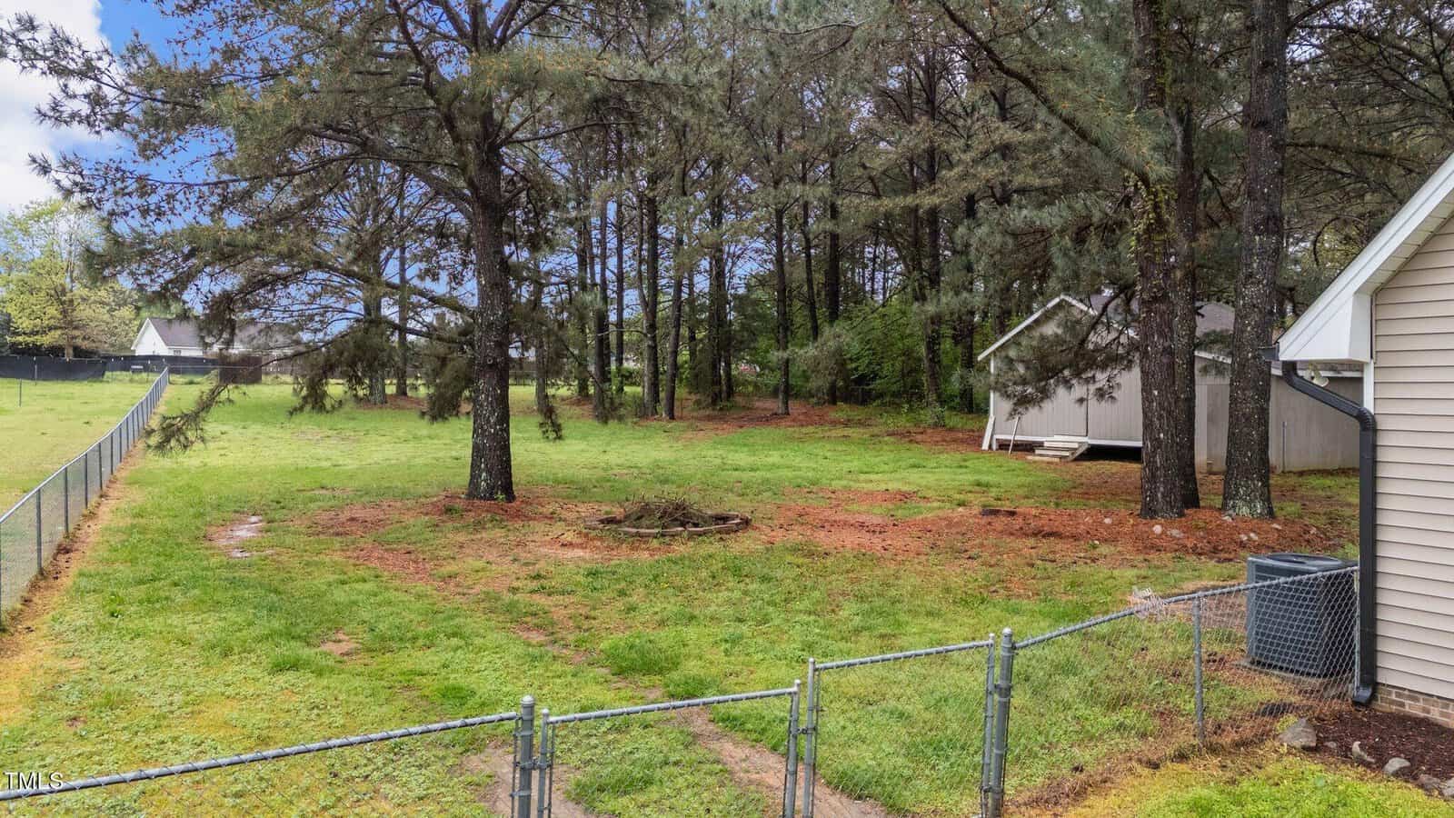 Fenced backyard with several pine trees, a small patch of bare ground, and a detached shed in the background. House siding and an air conditioning unit are visible on the right side. Grass covers most of the yard.