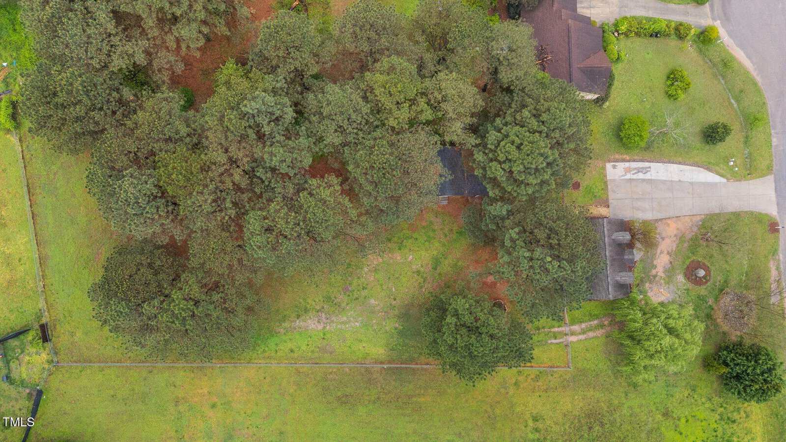 Aerial view of a property with dense trees covering most of the area, a driveway leading to a house partially covered by trees, and neighboring yards with grass and shrubs visible.