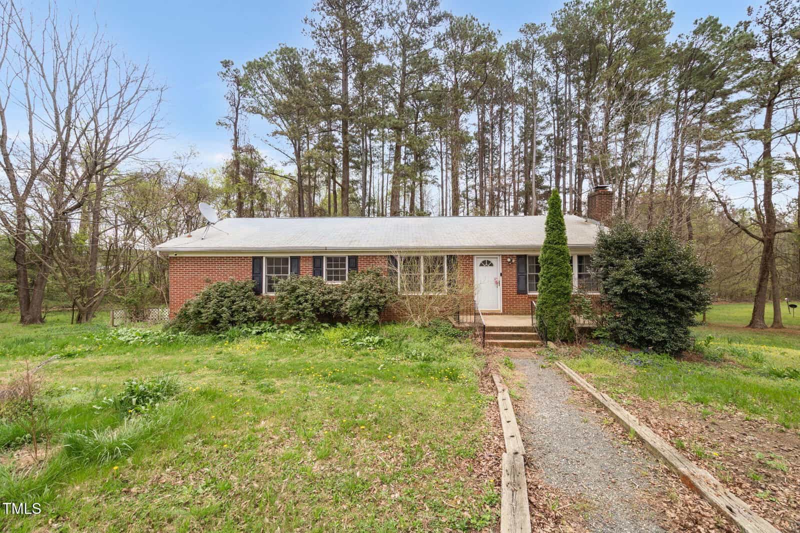 A brick house on 6702 Huntsboro Road, Oxford, with a white roof, stands before a dense forest. The yard is overgrown with grass and shrubs. A narrow gravel path leads to the entrance featuring a wooden door and small front porch. Tall trees loom behind the house.