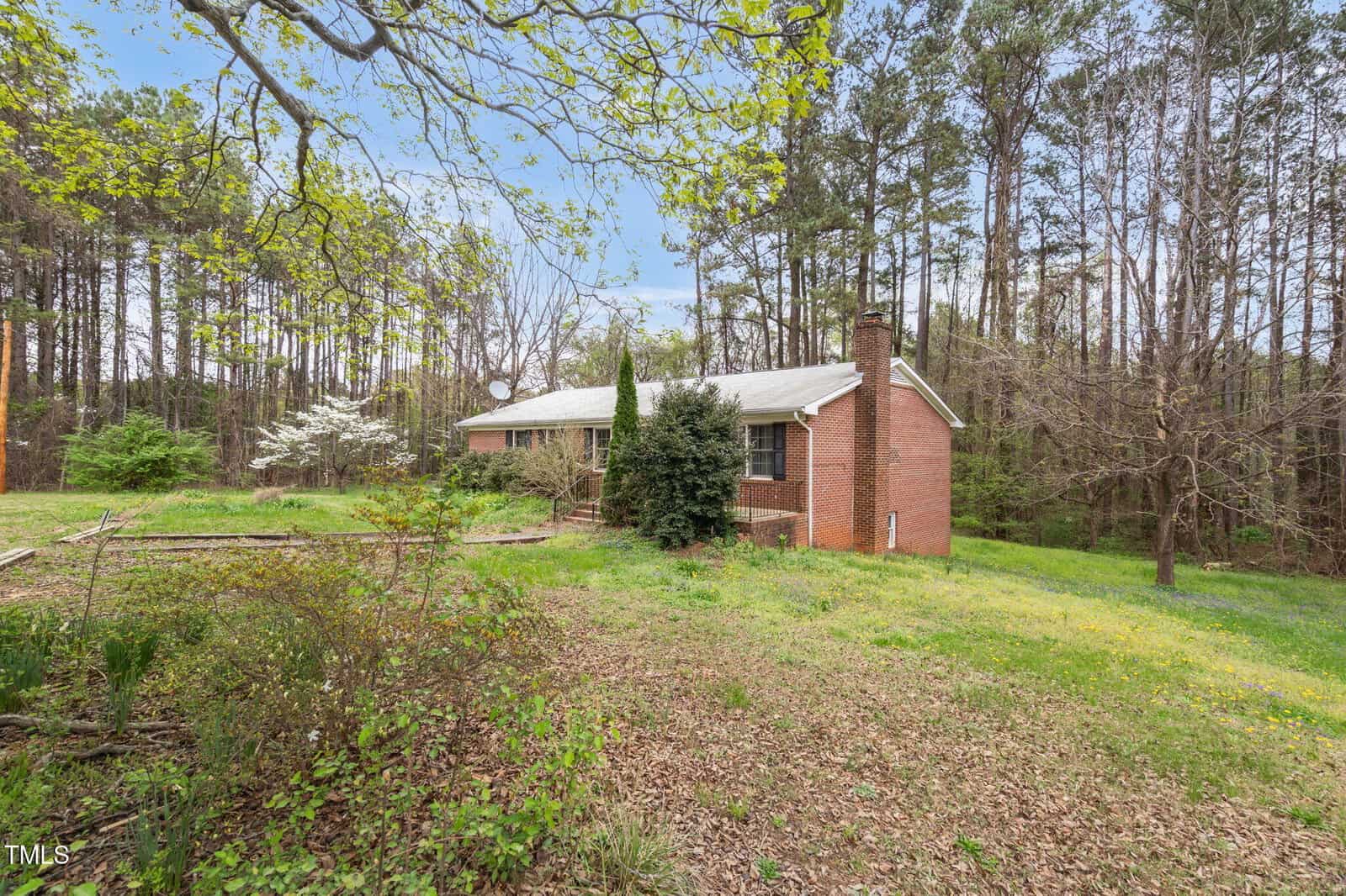 A brick house with a chimney is situated in a wooded area, surrounded by trees and a grassy yard. The house has a single story and a sloped roof. Leafless trees and some green shrubs are visible near the house.