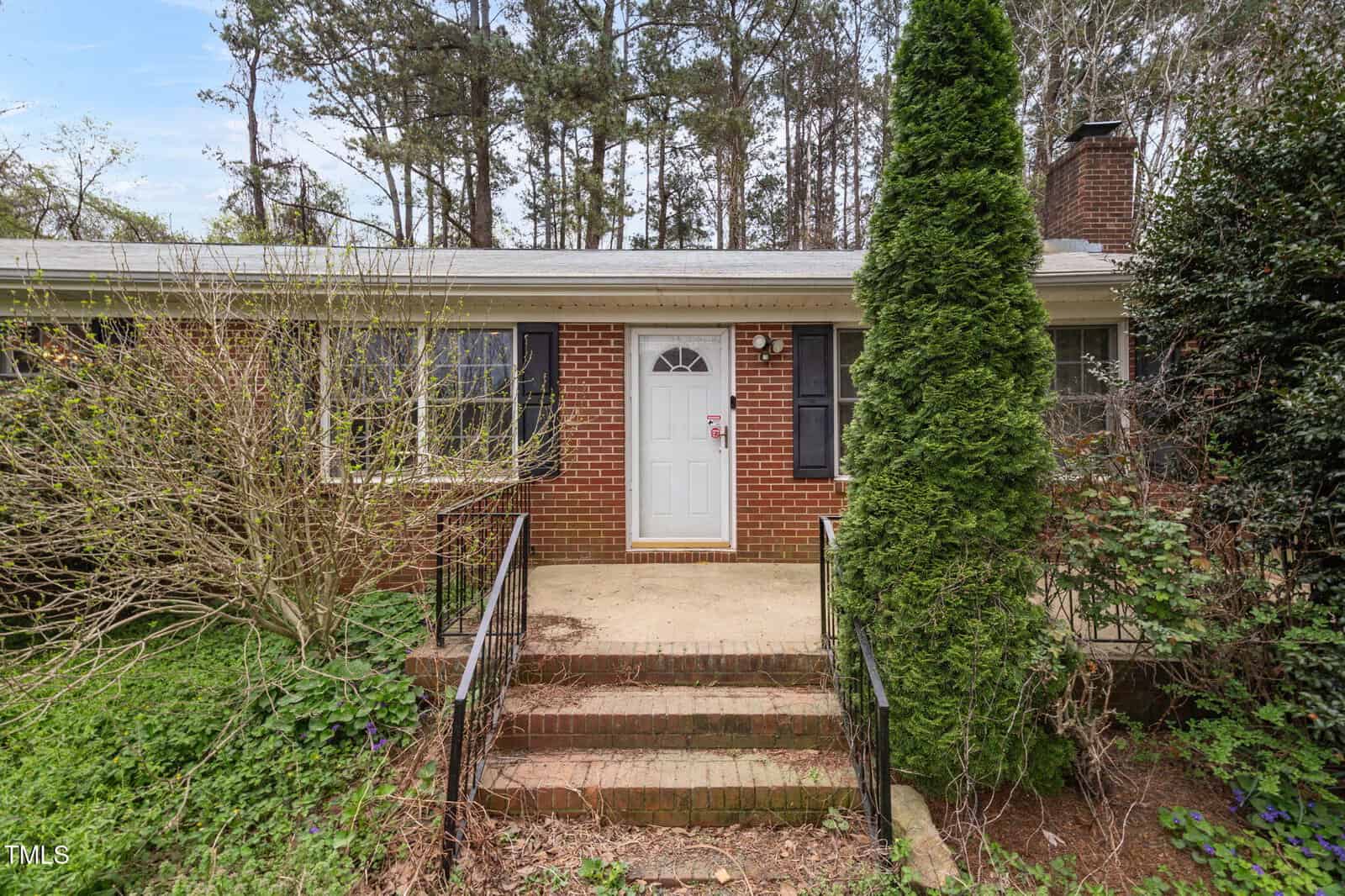 A brick house with a white door, flanked by large windows and a slender evergreen tree on the right. Steps lead up to the entrance, surrounded by dense, overgrown foliage. Tall trees are visible in the background.