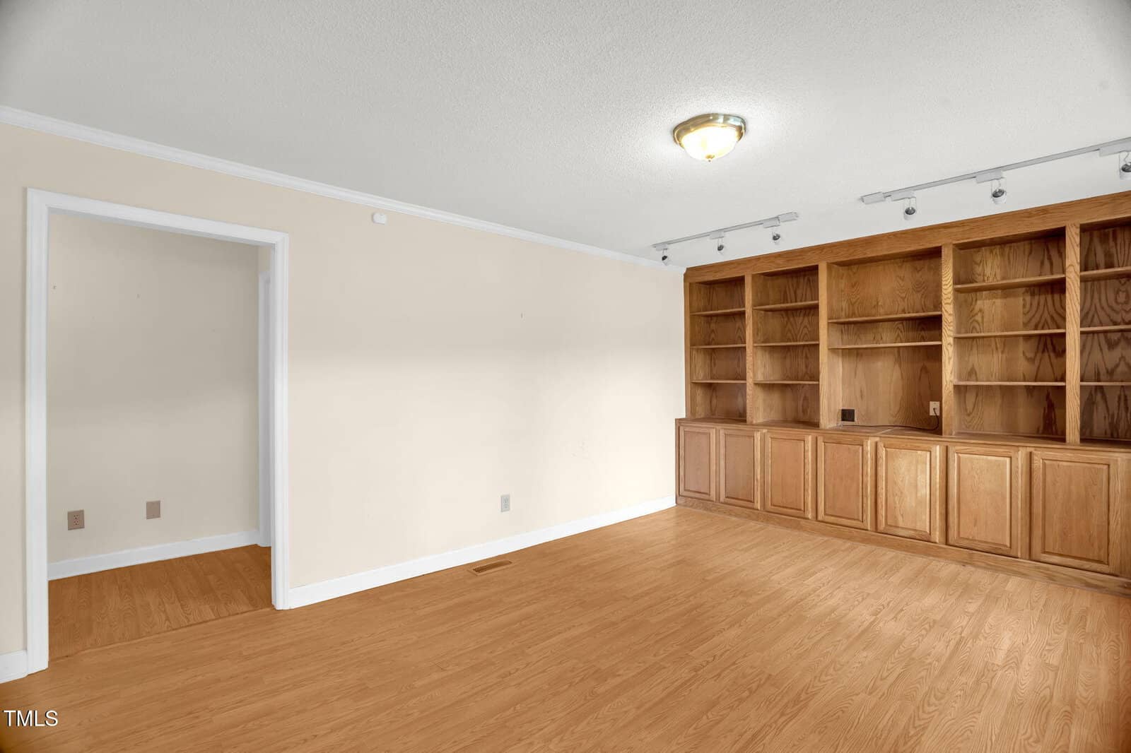 A room with light-colored walls and a wooden floor features built-in wooden shelves and cabinets along one wall. There is a ceiling light fixture and a doorway leading to another room. The space is empty and well-lit.
