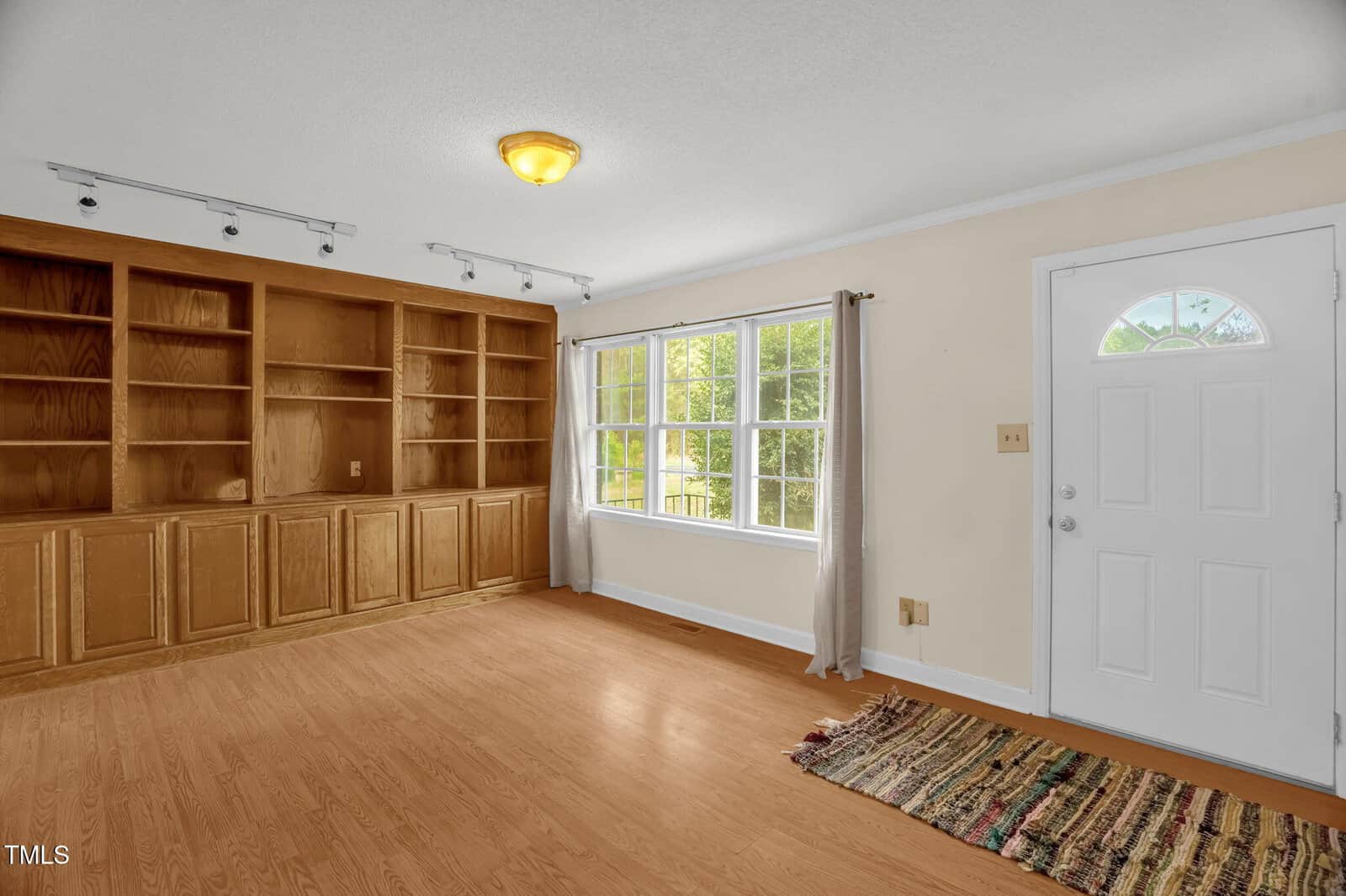 A room with light wood flooring and beige walls features built-in wooden shelves on the left. A large window with a curtain is on the right, next to a white door. A ceiling light fixture and a patterned rug are visible.