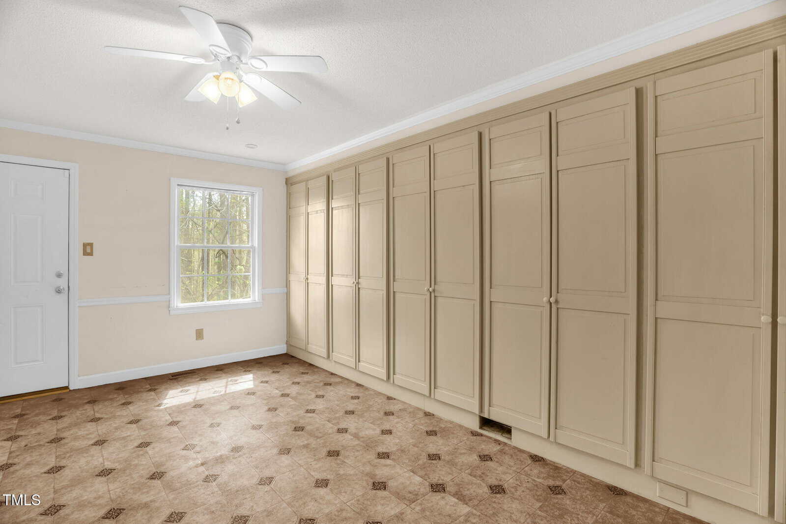 Room with a tiled floor, ceiling fan, and beige wall-to-ceiling cabinets on the right. A window is on the left, allowing natural light in. A white door is in the back left corner. The walls are cream-colored with a white trim.