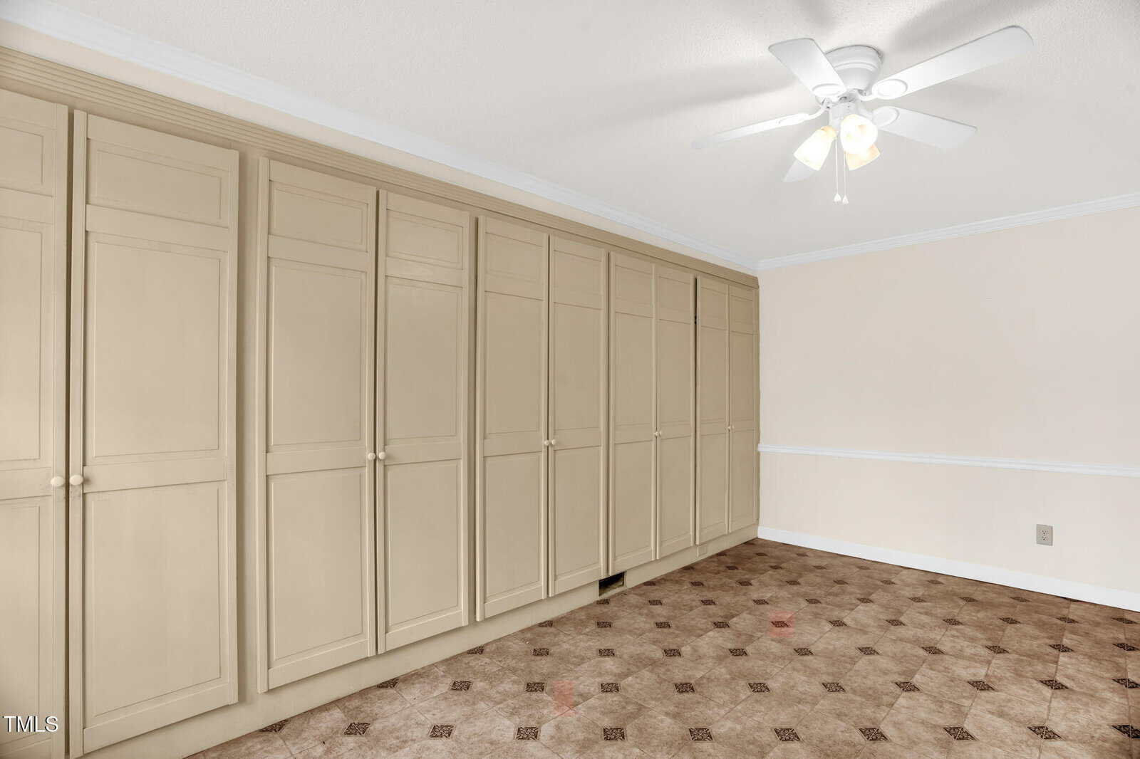 A room with a row of beige built-in cabinets along one wall, a ceiling fan with a light fixture, and patterned carpet flooring. The walls are plain with a chair rail trim. The space is well-lit with natural light.