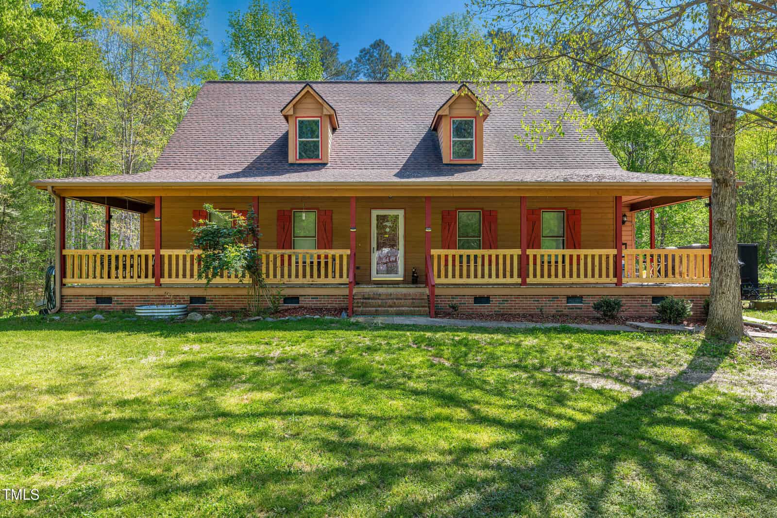 A wooden house with a large covered front porch and dormer windows sits on a grassy lawn, surrounded by trees and greenery under a clear sky.