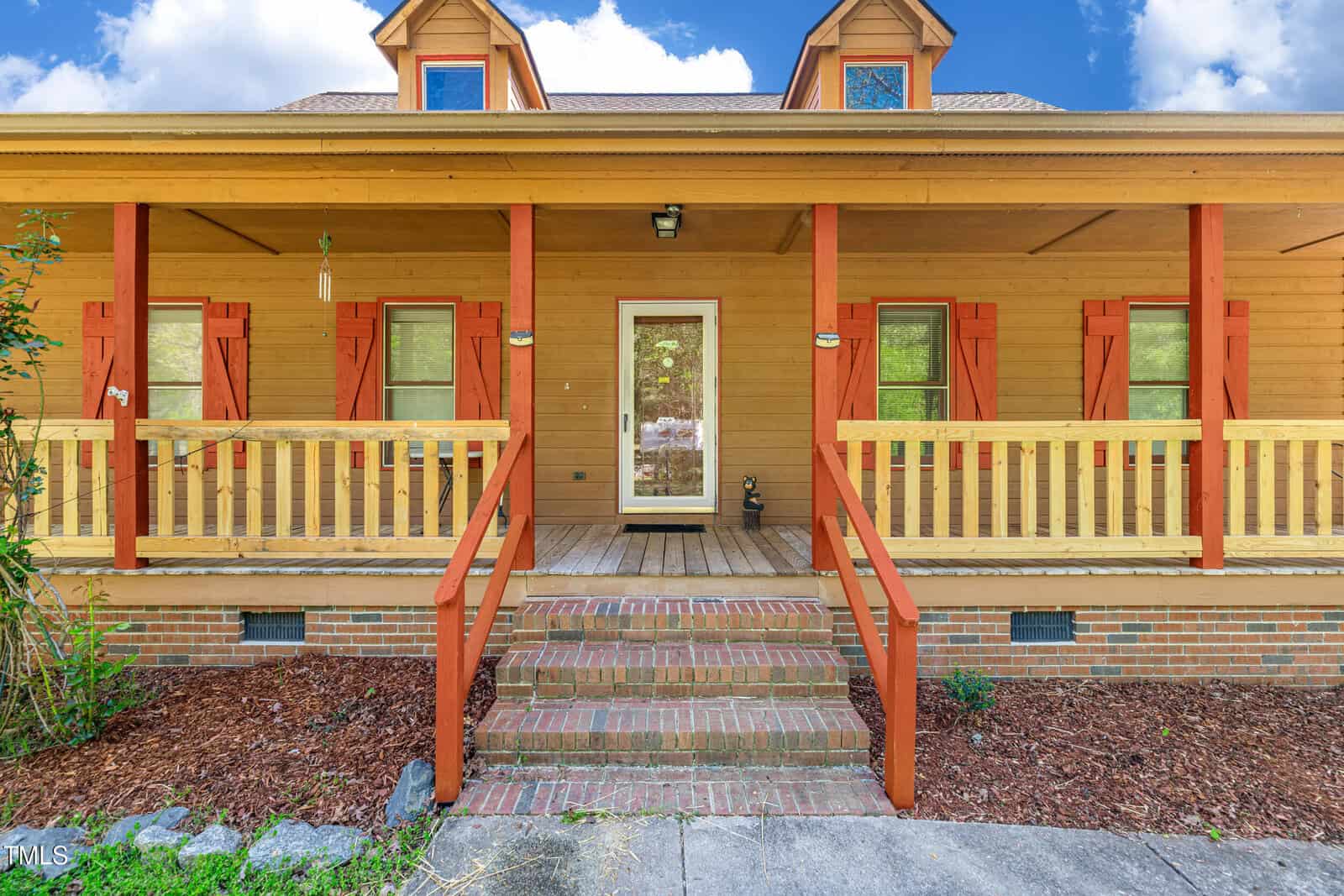 Front view of a wooden house with a brick porch, two sets of stairs, red shutters, and a covered front porch with a yellow railing. There are two dormer windows on the roof and landscaping along the walkway.