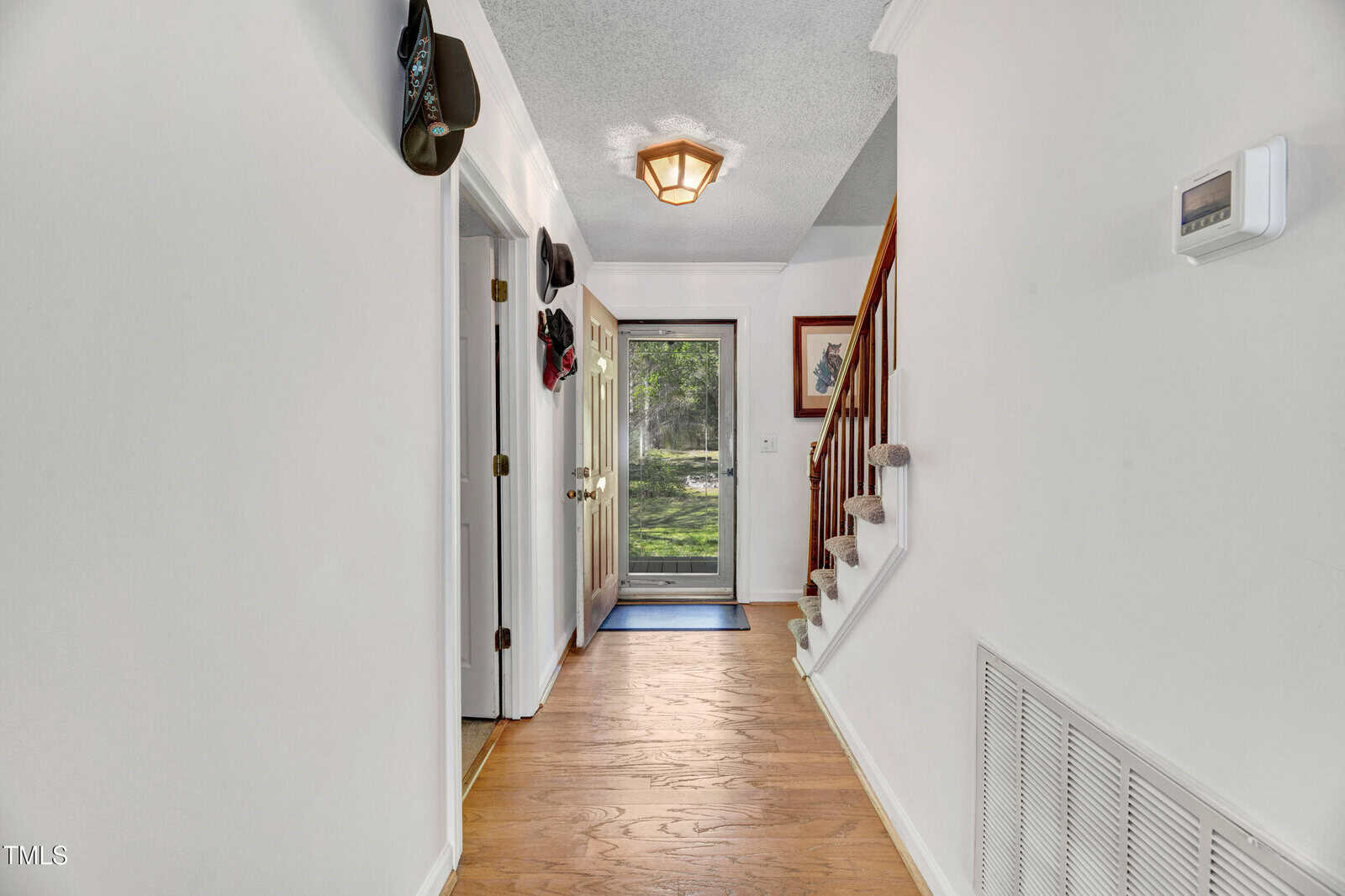 A hallway with hardwood floors, white walls, a staircase with carpeted steps, hats hanging on wall hooks, open doors to the left, and a glass door at the end leading outside.