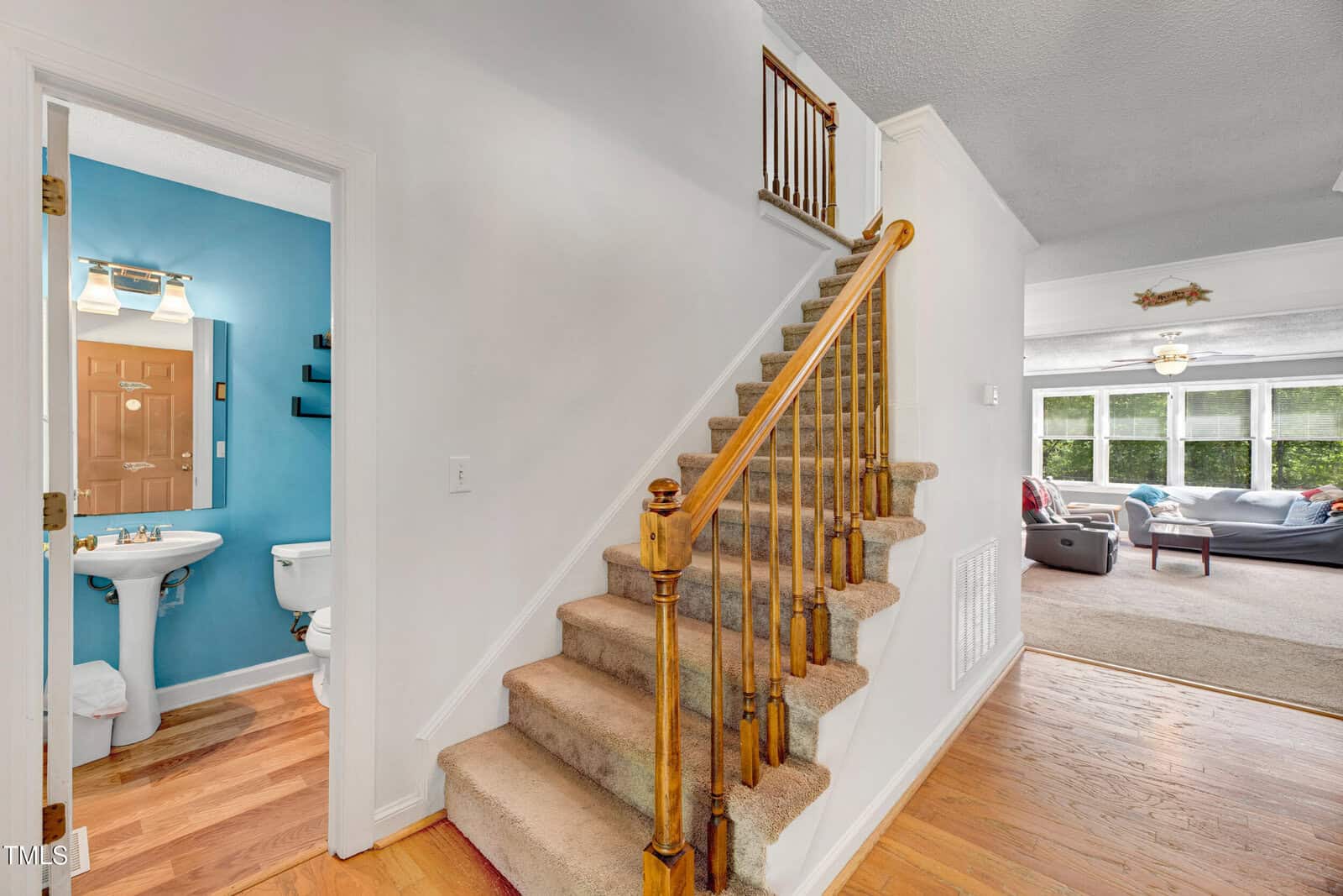 A carpeted staircase with a wooden banister is beside a hallway. To the left is a bathroom with blue walls, a pedestal sink, and a toilet. Straight ahead is a living room with large windows and seating.