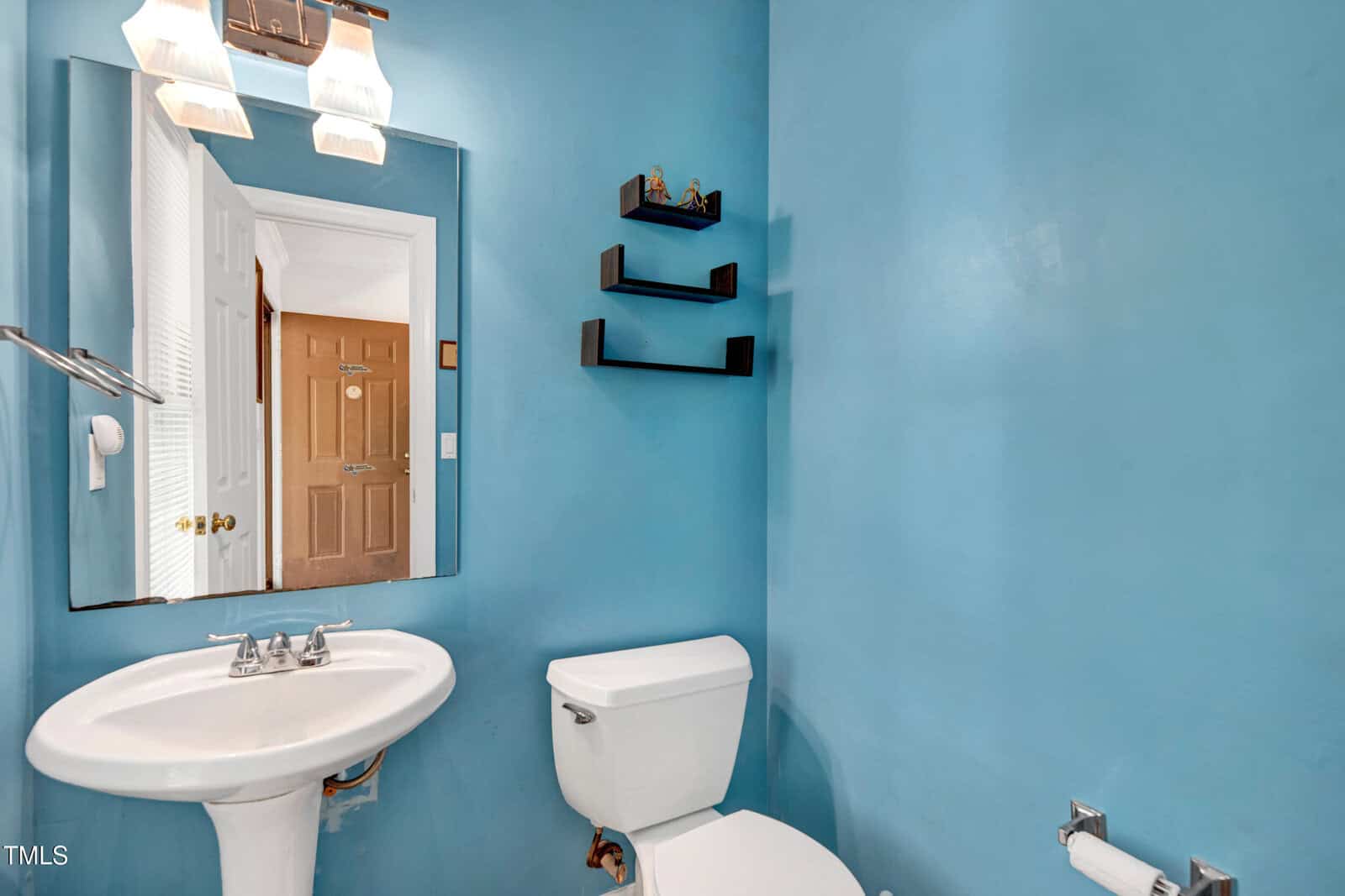 A bathroom with blue walls featuring a pedestal sink, a toilet, a wall mirror, three small black shelves, and a towel holder. The door is open, revealing a wooden door in the hallway beyond.