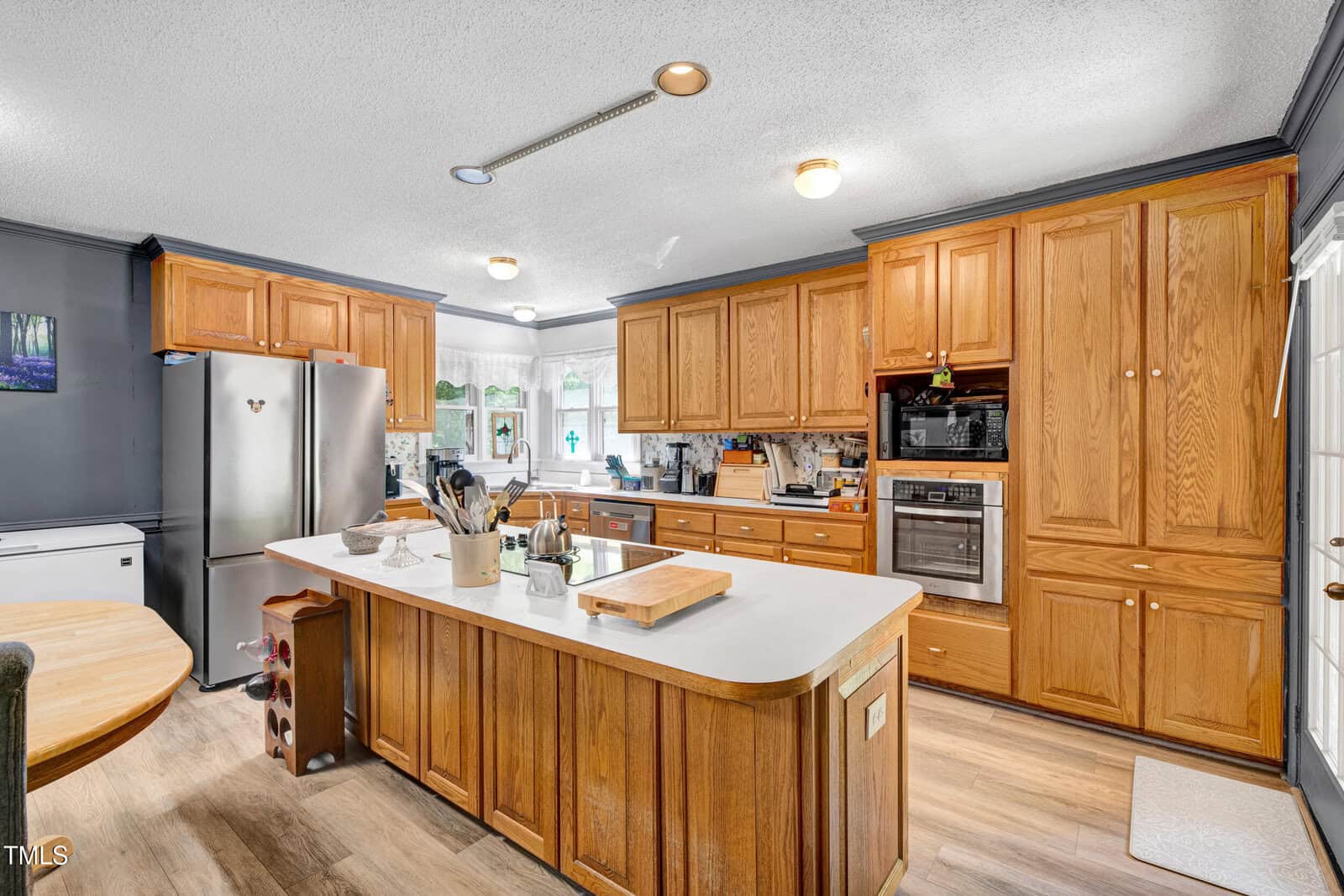 A kitchen with wooden cabinets, a center island with a white countertop, stainless steel refrigerator, built-in oven, various kitchen utensils, and a window above the sink letting in natural light.