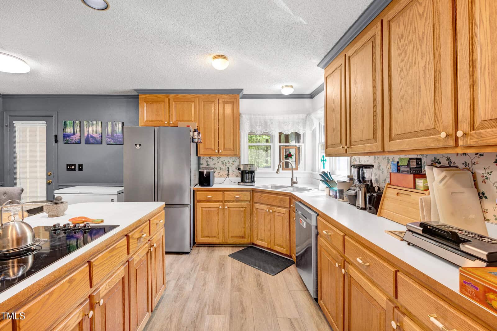 A kitchen with wooden cabinets, white countertops, stainless steel appliances, a stove, a refrigerator, a dishwasher, small kitchen appliances, and a window above the sink with white curtains.