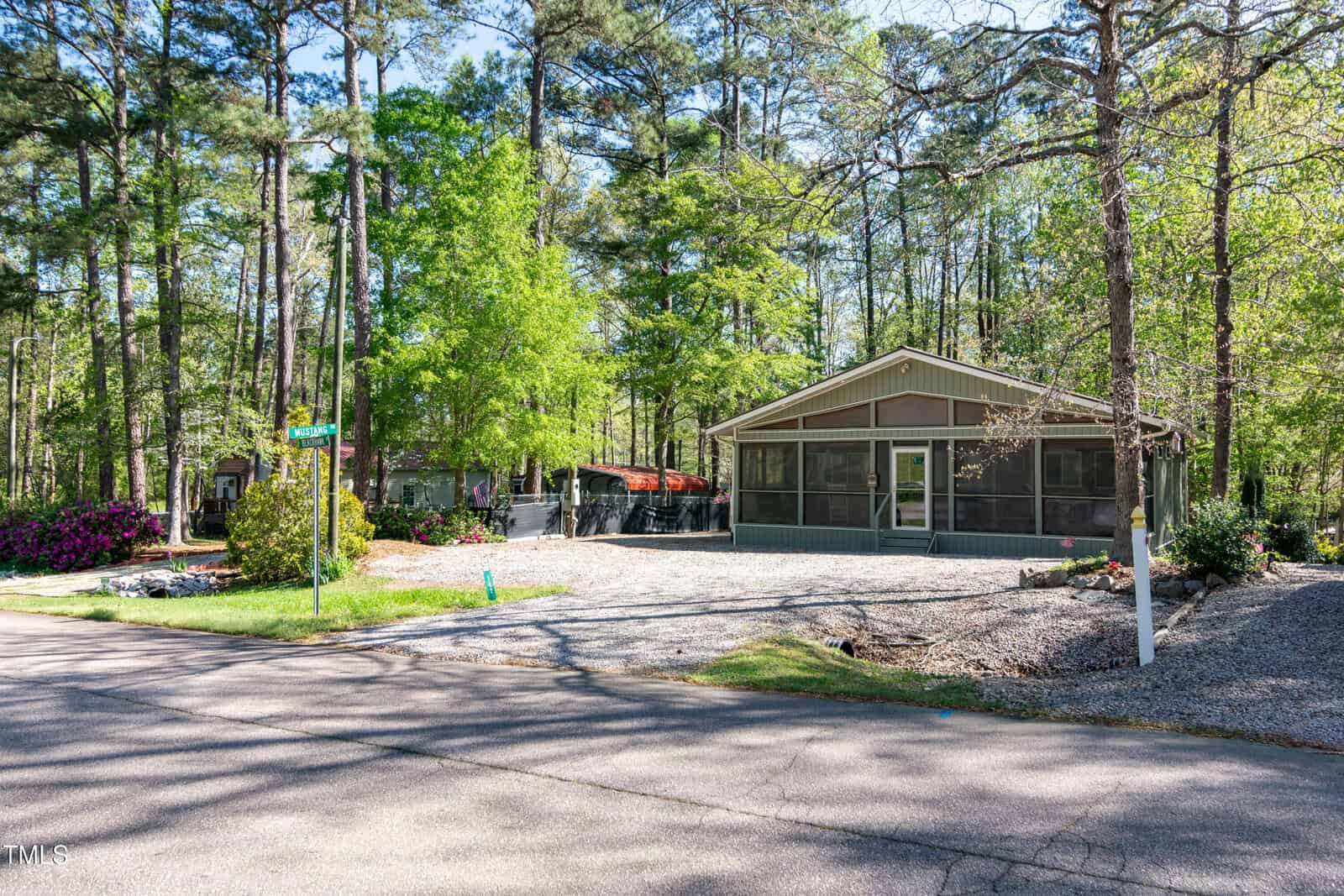 Single-story house with screened porch sits among tall trees. The yard is covered with gravel and bordered by bushes and flowers. A quiet paved street runs in front, with a street sign visible on the corner.