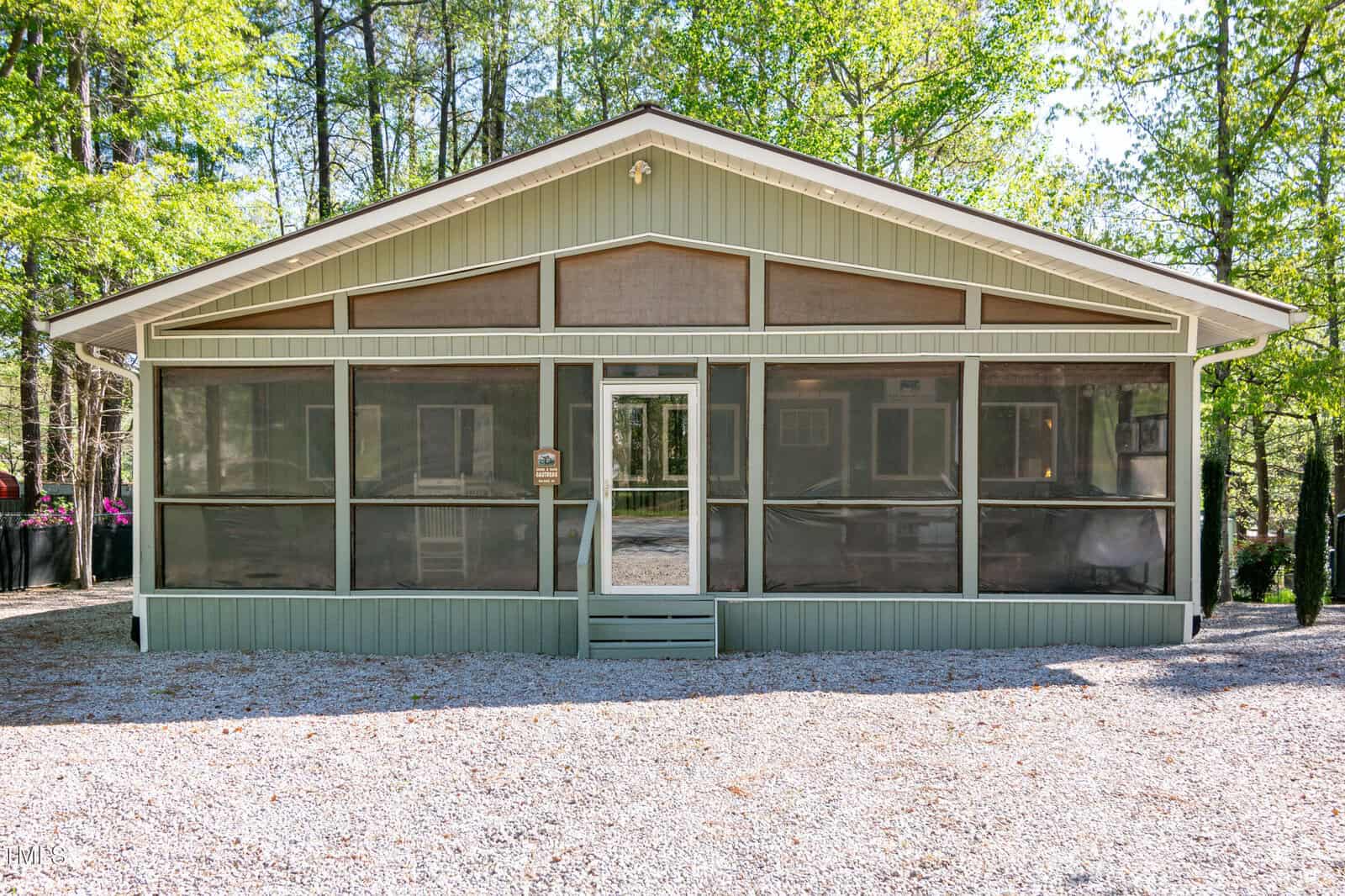 A single-story house with light green siding and a screened-in front porch, set on a gravel lot and surrounded by tall trees in a wooded area.