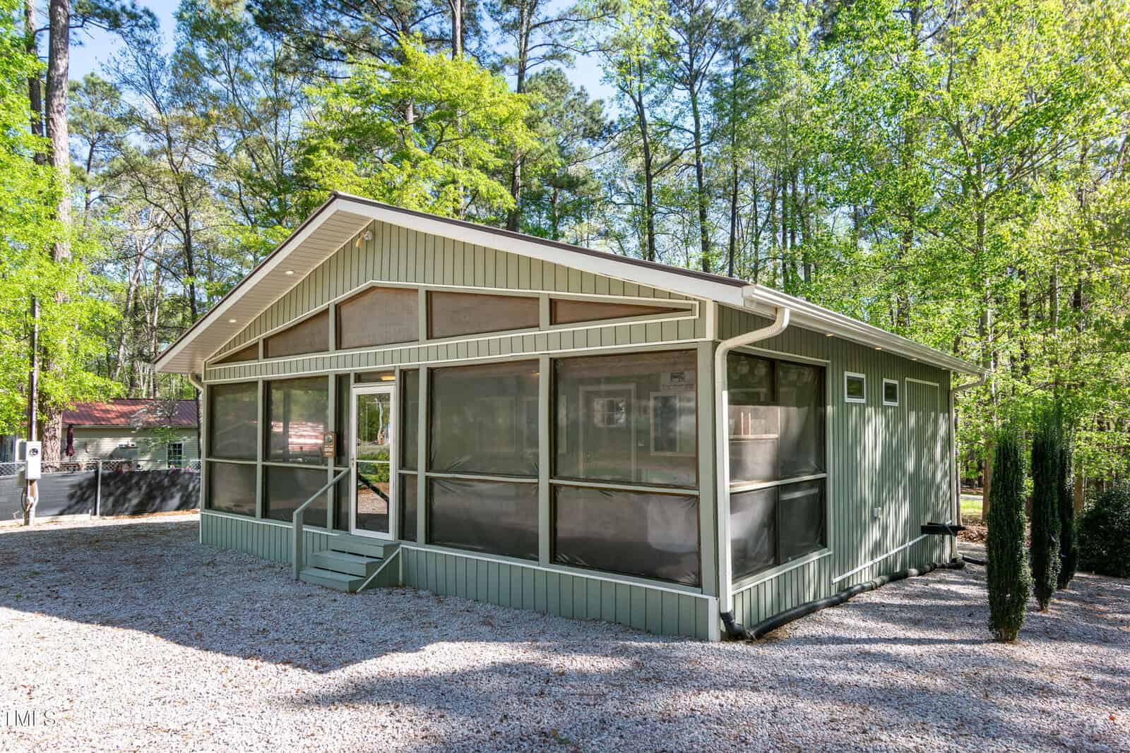 A single-story green house with a screened-in porch sits on a gravel lot, surrounded by tall trees and greenery under a clear sky.