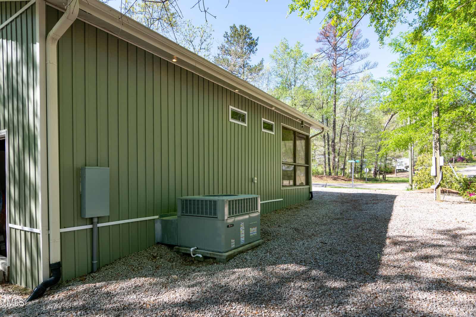 A large green metal-sided building with multiple narrow windows sits on a gravel lot. An air conditioning unit and utility box are positioned against the exterior wall. Trees and a road are visible in the background.