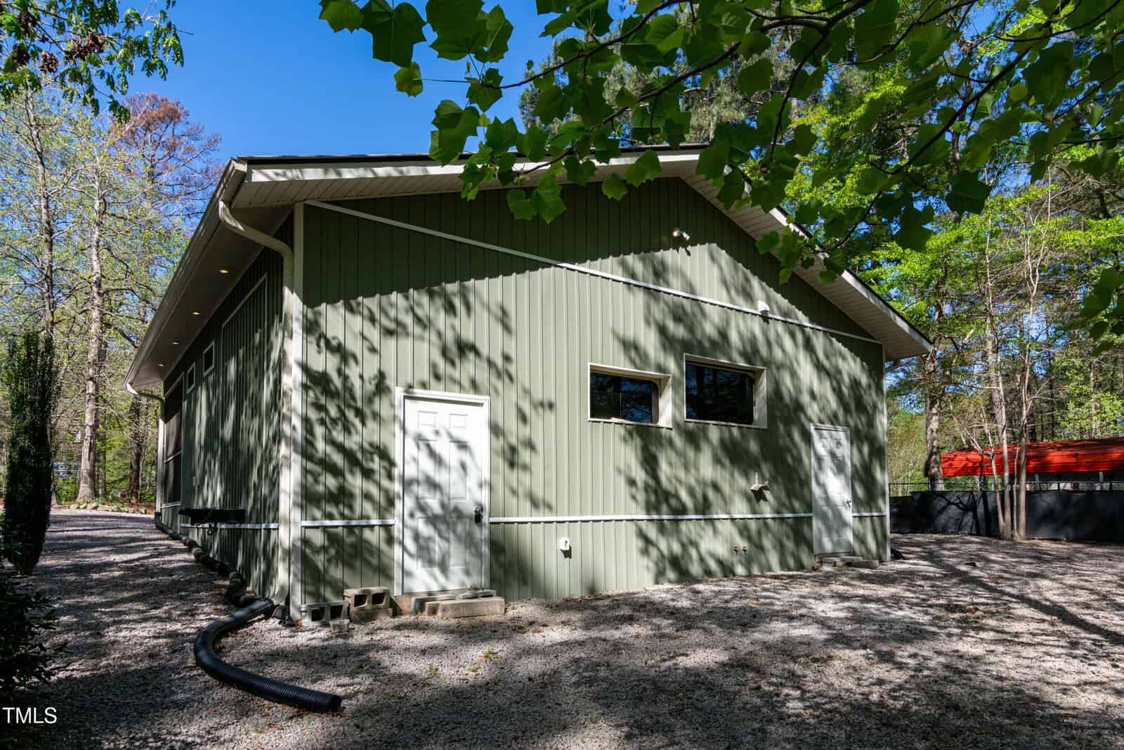 A large green metal building with a white door, two rectangular windows, and a gravel driveway surrounded by trees and greenery on a sunny day.