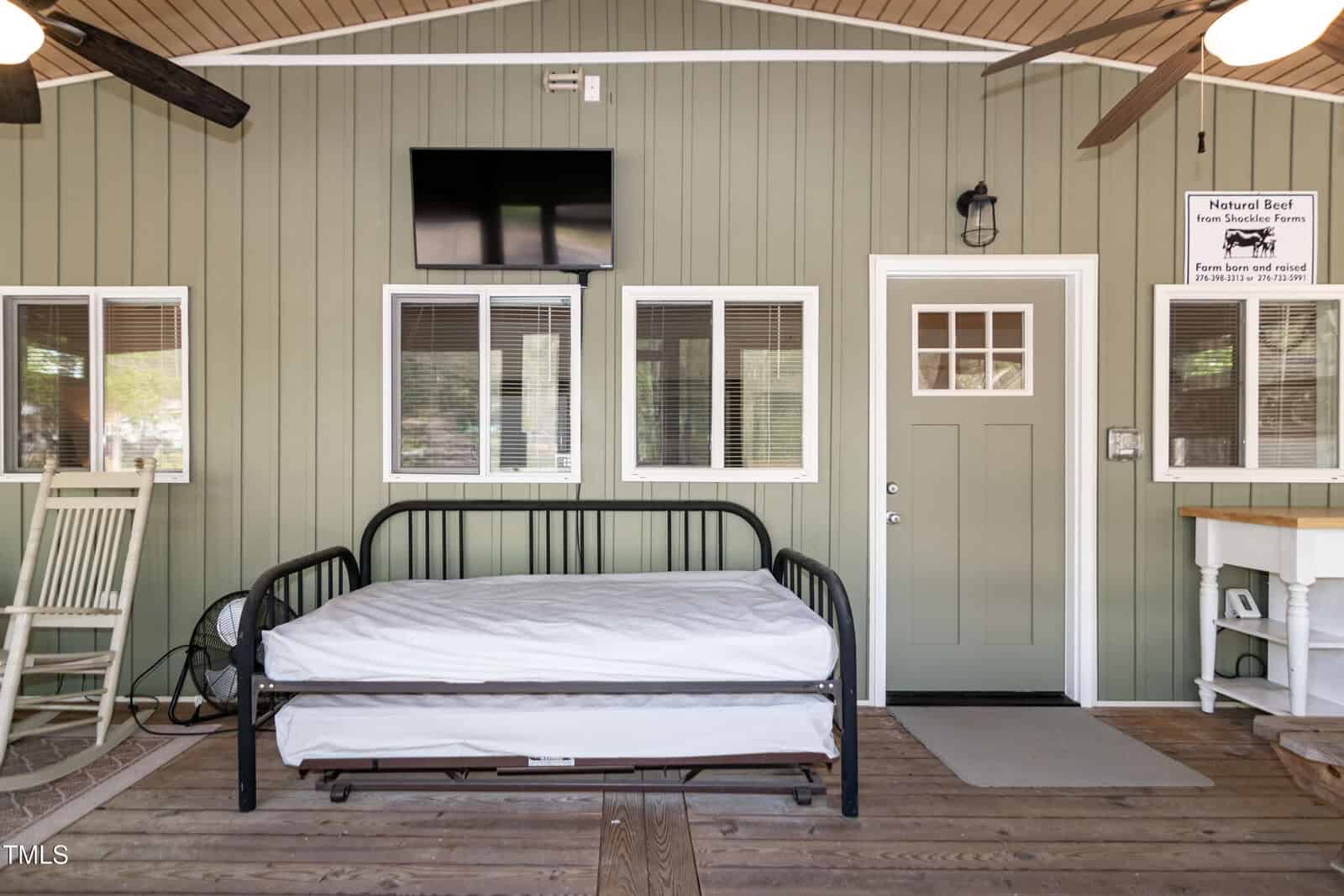 A simple room with a metal bed frame and mattress, rocking chair, TV mounted on a green paneled wall, several windows, a door, ceiling fan, and a small counter with a farm sign above it.