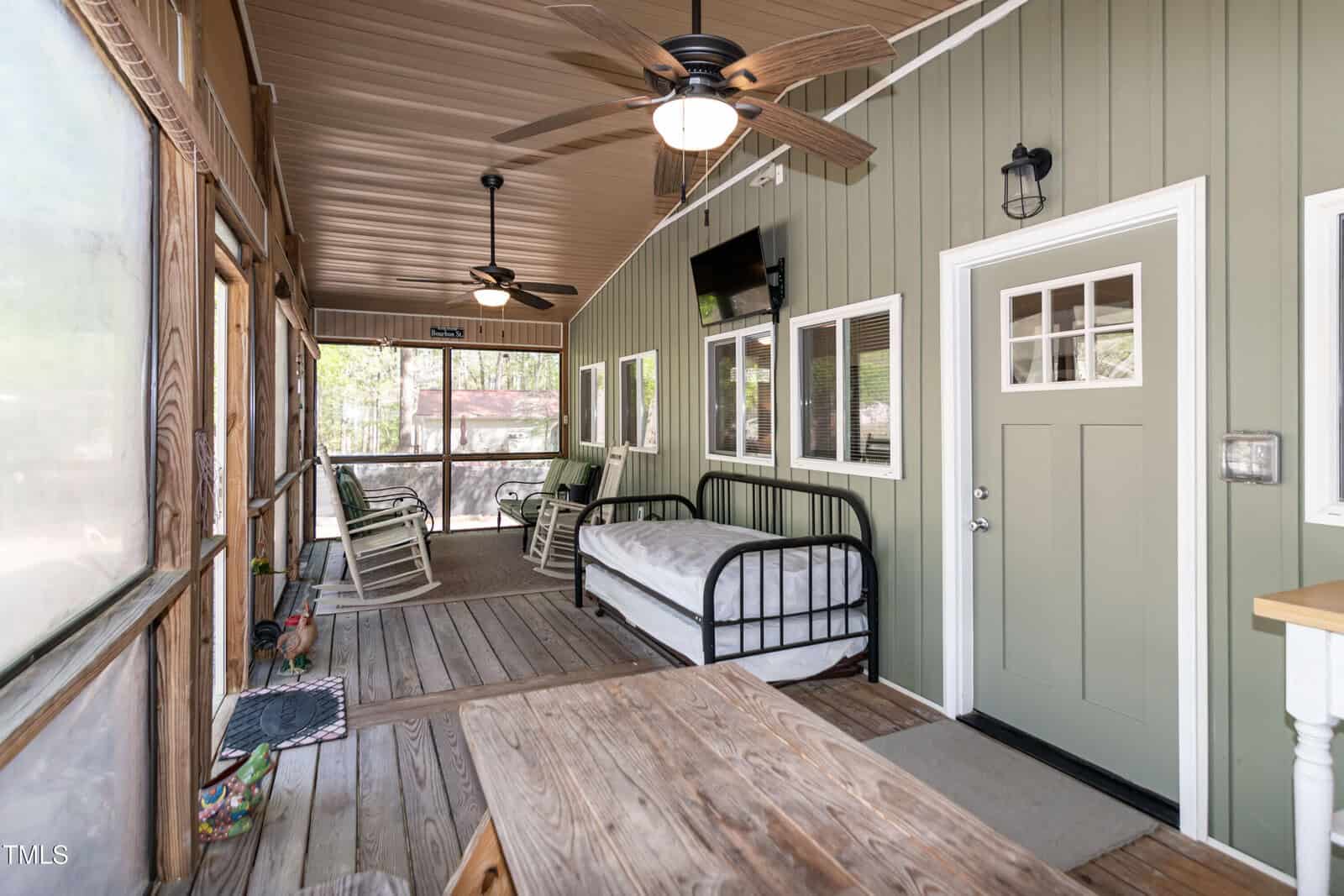 Screened porch with a ceiling fan, rocking chairs, a daybed, wooden dining table, and green siding walls. Multiple windows, a wall-mounted TV, and a door with glass panels are visible. Natural light enters from the outside.