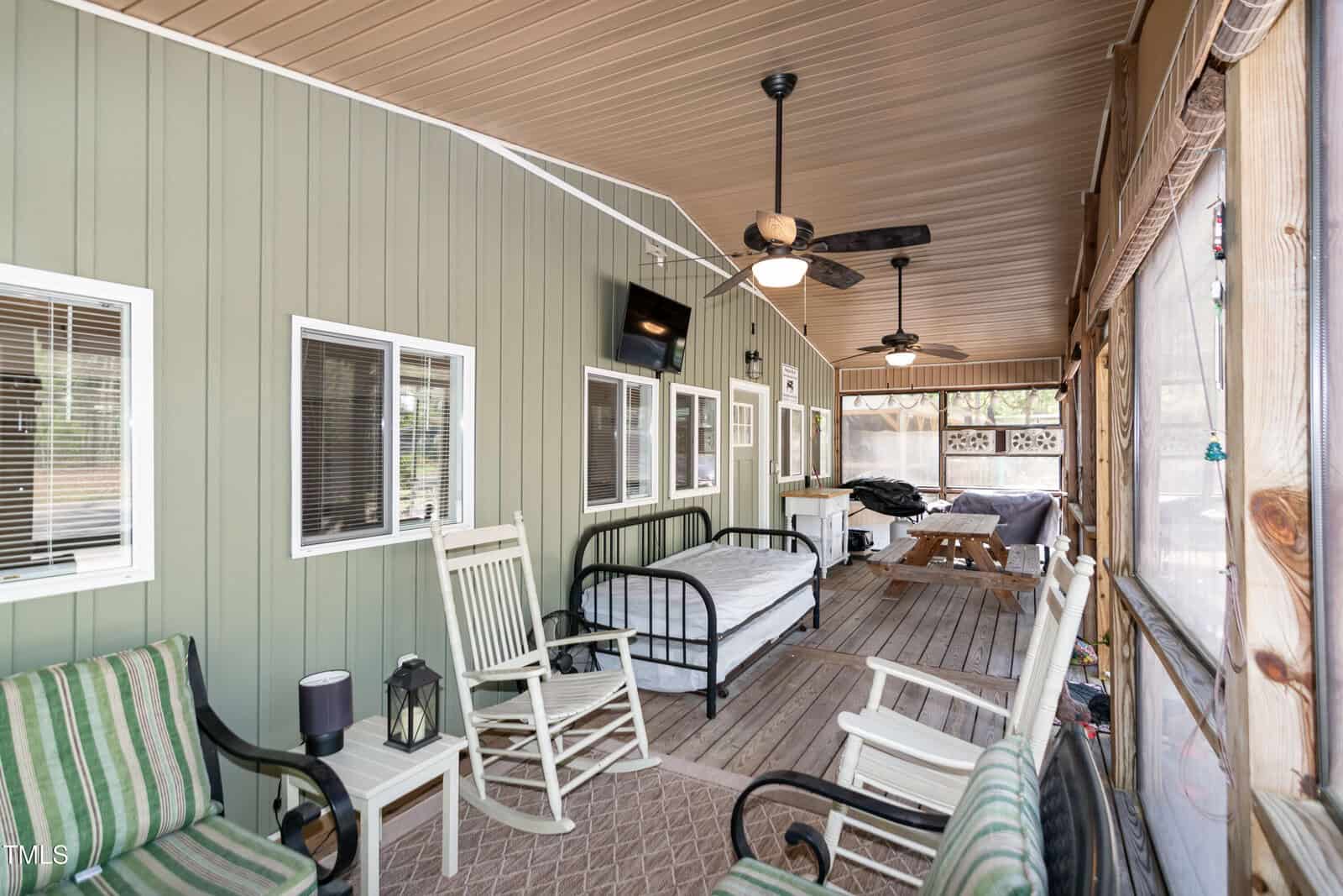 Screened porch with green siding, rocking chairs, a bed, a bench, ceiling fans, picnic table, and a rug. Windows line the wall, and the porch overlooks an outdoor area, creating a casual, multi-use space.