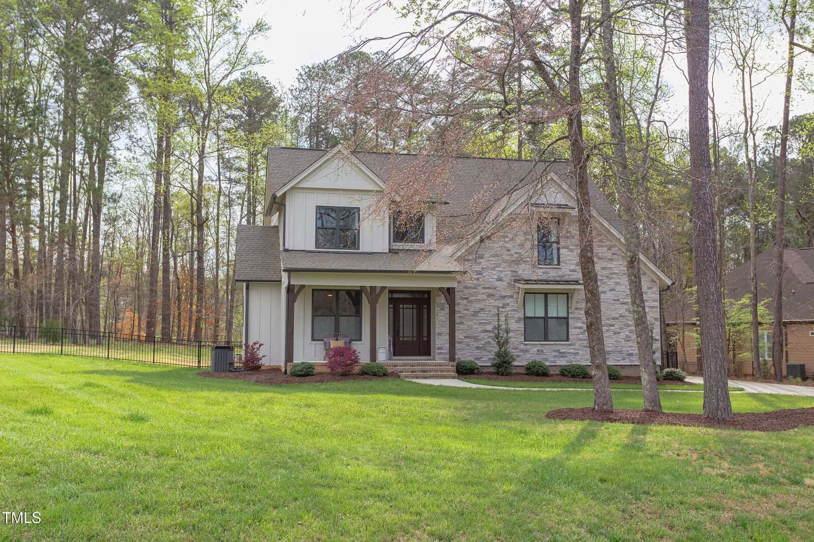 A two-story house with stone and siding exterior stands on a well-maintained lawn, surrounded by trees. The house has multiple windows and a gabled roof. A paved walkway leads to the covered entrance, and a fence is visible in the background.