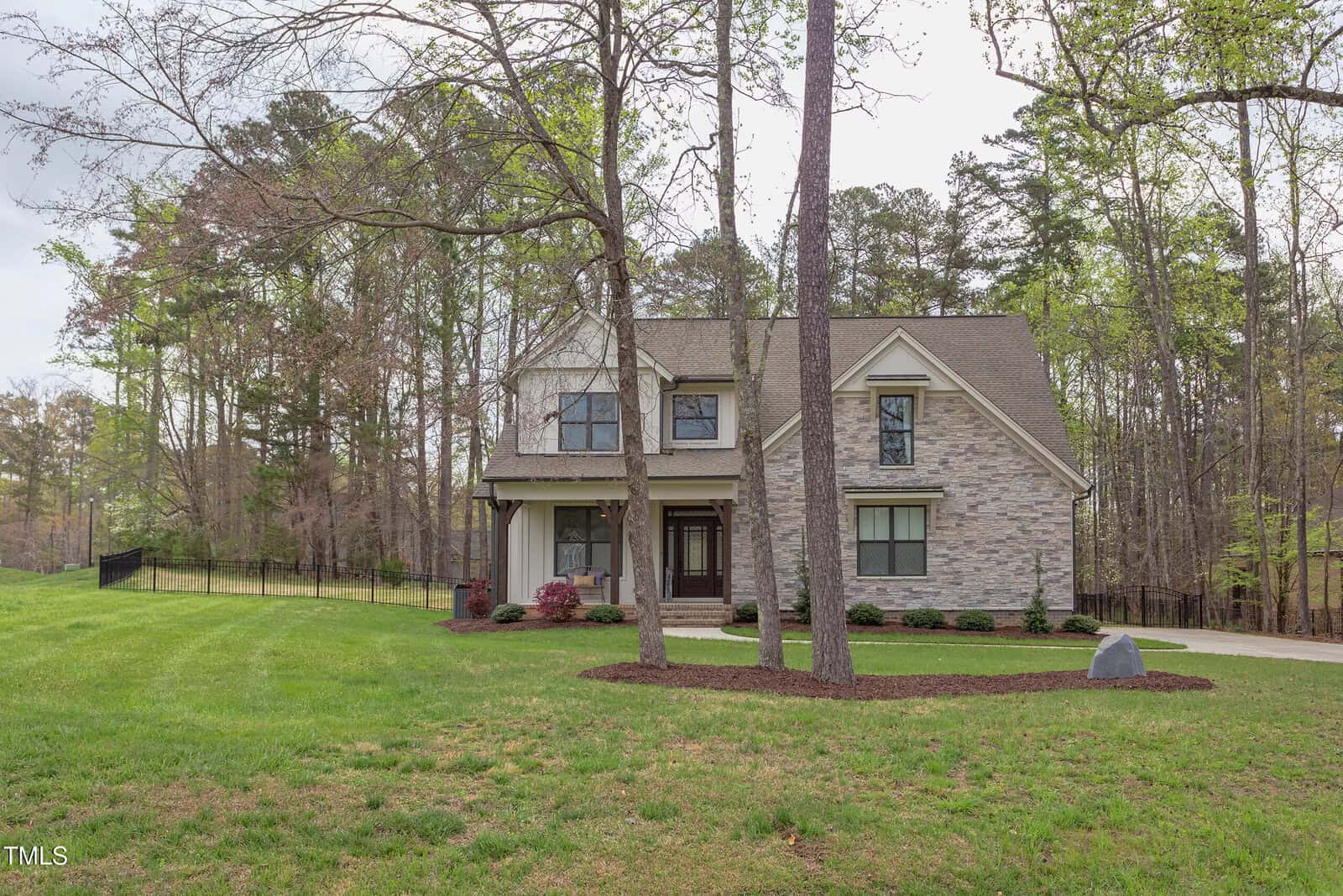 Two-story house with stone and beige siding, large front windows, and a covered entryway. It is surrounded by a green lawn and tall trees, including a few in the front yard. A driveway leads to the right side of the house.