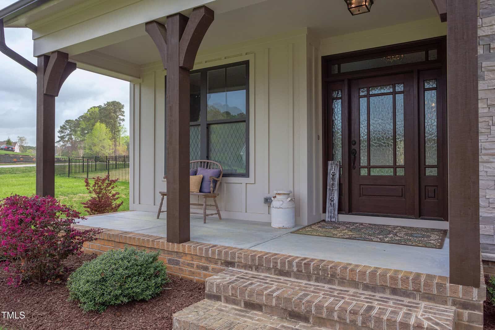 Front porch of a house with wooden columns and a brick step. There is a dark wooden door with glass panels, a small window, a brown chair with a cushion, a white milk canister, and a patterned doormat. Bushes and trees are visible in the background.