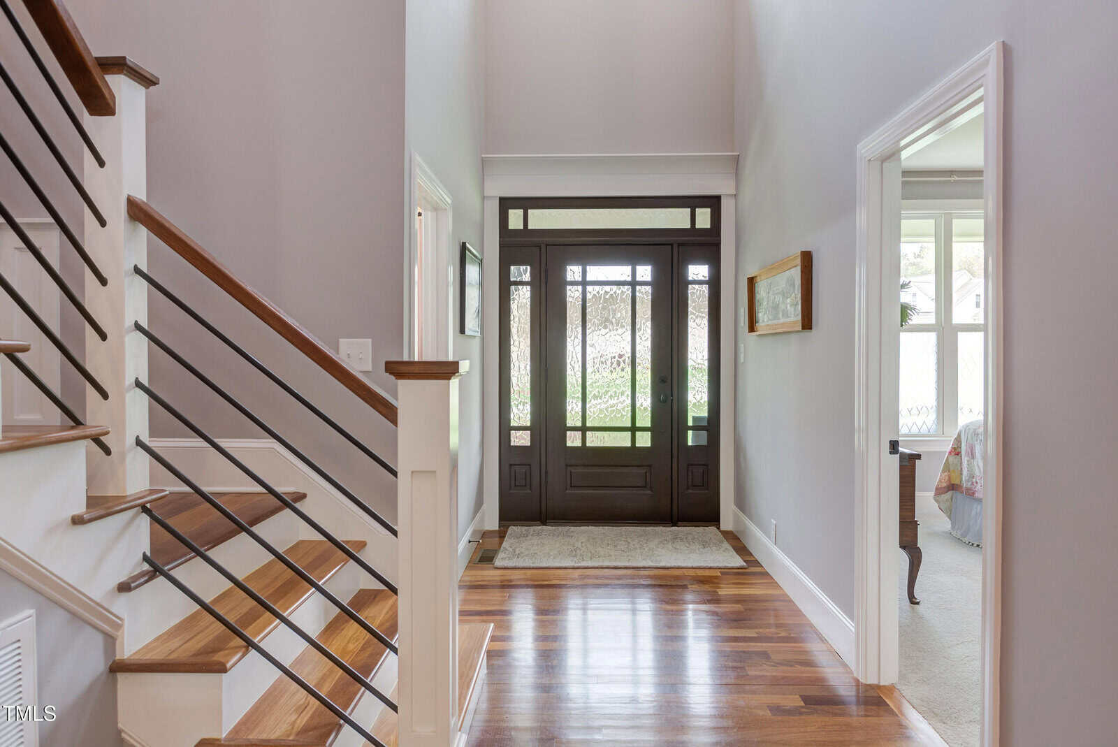 A bright foyer with wooden flooring, featuring a staircase on the left with modern railing. A dark wooden front door with glass panels is in the background. The foyer connects to other rooms through open doorways on the right.