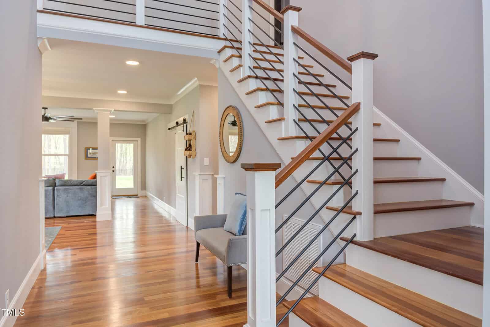 A modern home interior featuring a wooden staircase with metal railings. The hallway has hardwood floors, a wall mirror, and a small bench with cushions. A doorway leads to a living area with a couch and large windows.