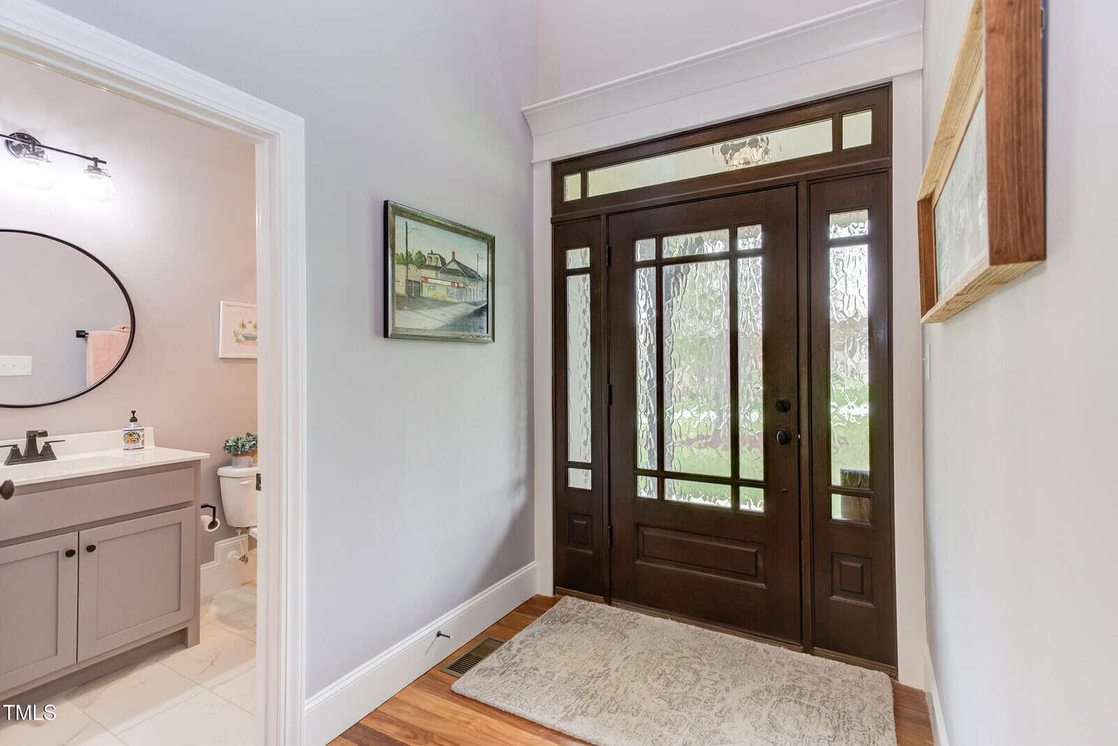 A hallway featuring a dark wooden front door with frosted glass panels. To the left is a doorway leading to a bathroom with a vanity and toilet visible. The walls are light-colored, and a framed artwork hangs above the door.