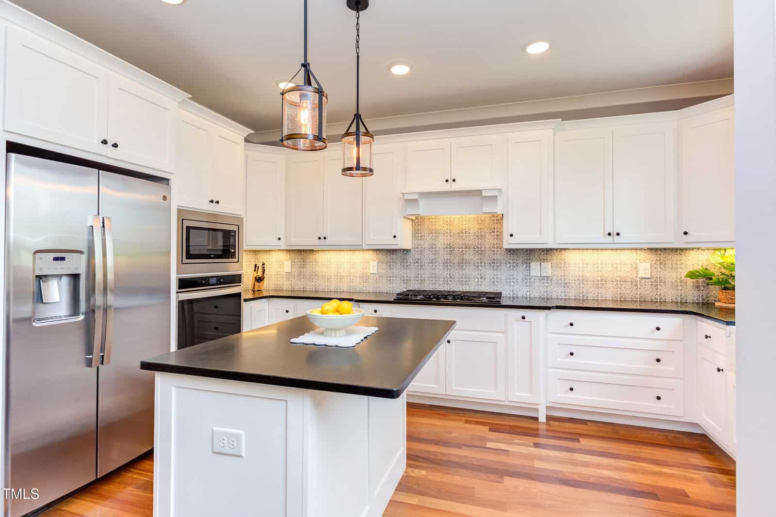 A modern kitchen features white cabinets with black countertops. A stainless steel refrigerator and built-in microwave are on the left. Two pendant lights hang above the central island, which holds a bowl of lemons. Wood flooring completes the space.