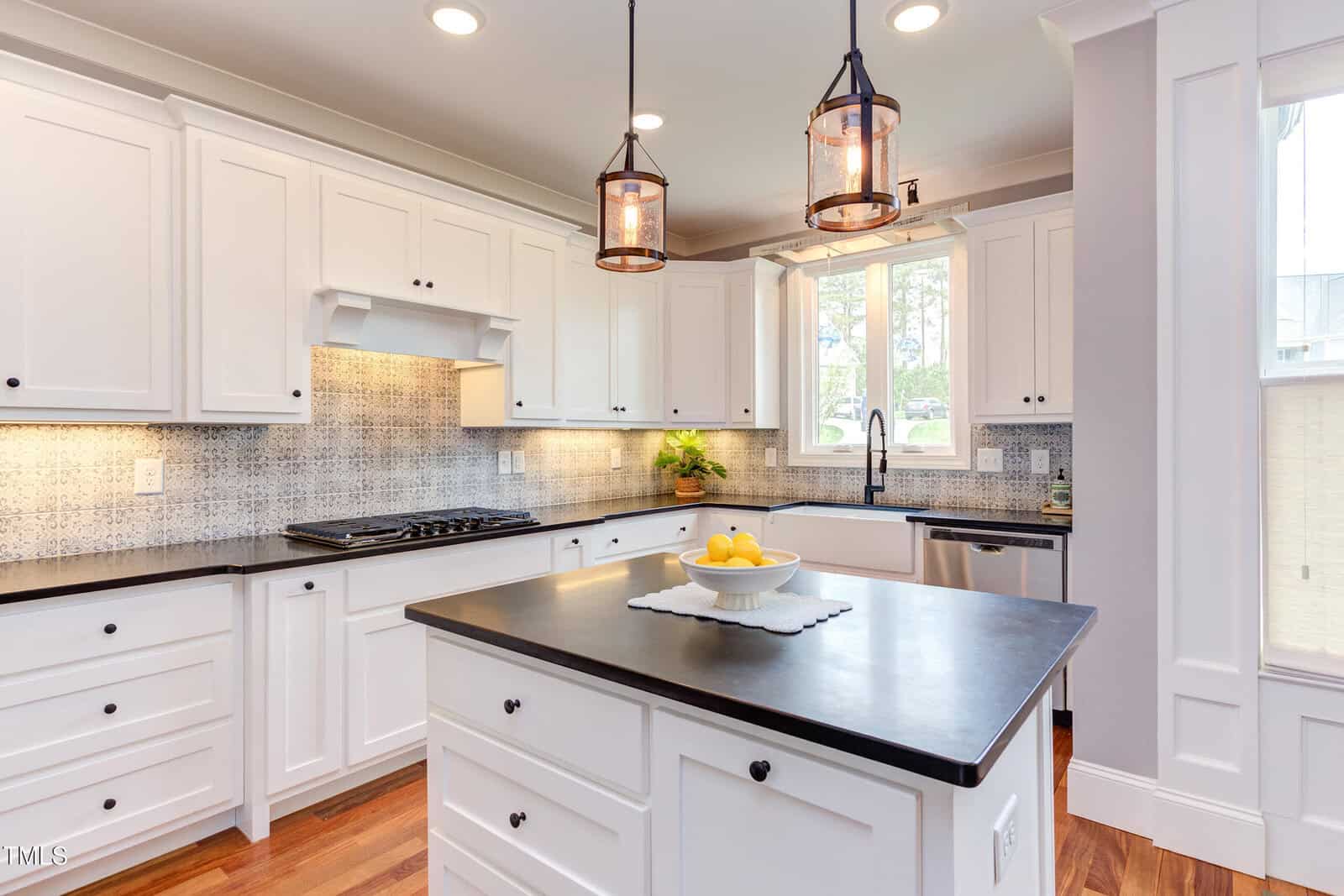 A modern kitchen with white cabinets, black countertops, and a central island. The backsplash features a patterned design. Two pendant lights hang above the island, and a bowl of lemons is placed on it. A window above the sink lets in natural light.