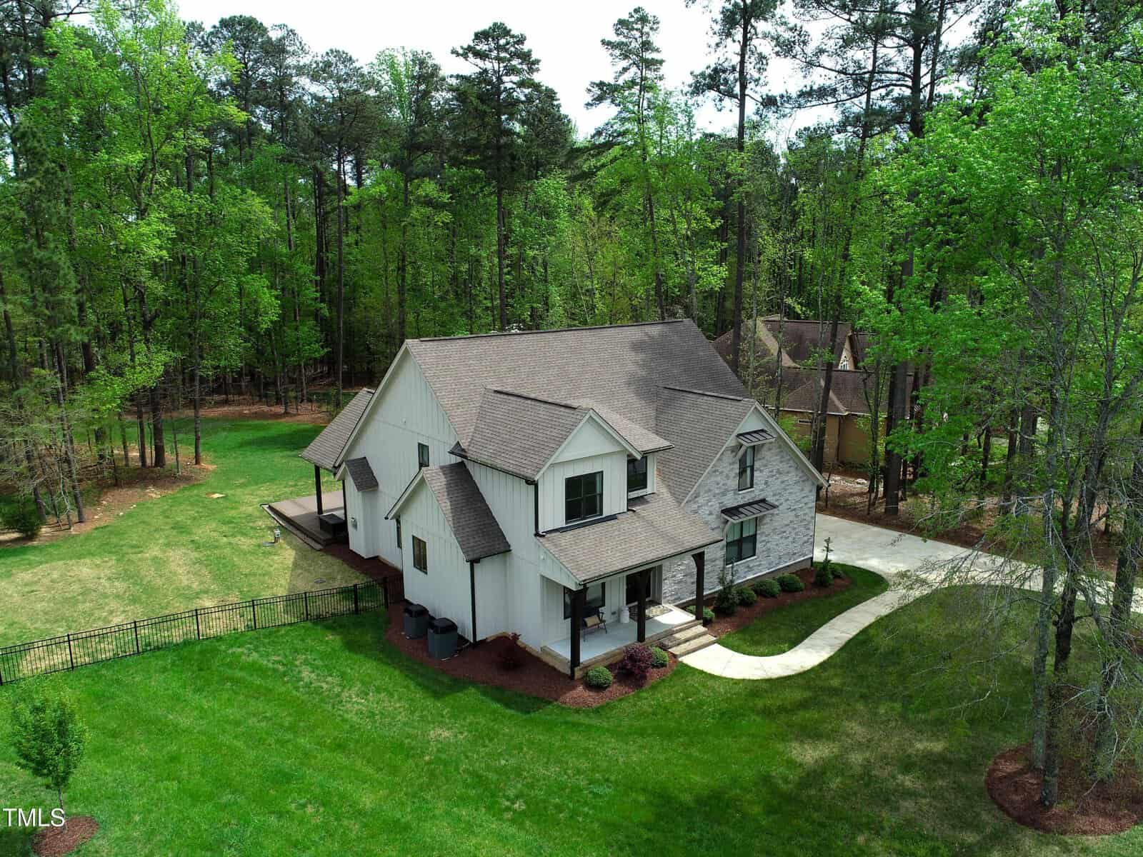 Aerial view of a two-story white house with a grey roof, surrounded by a large green lawn and trees. The property includes a driveway and a fenced backyard. The house features a covered front porch and several windows.
