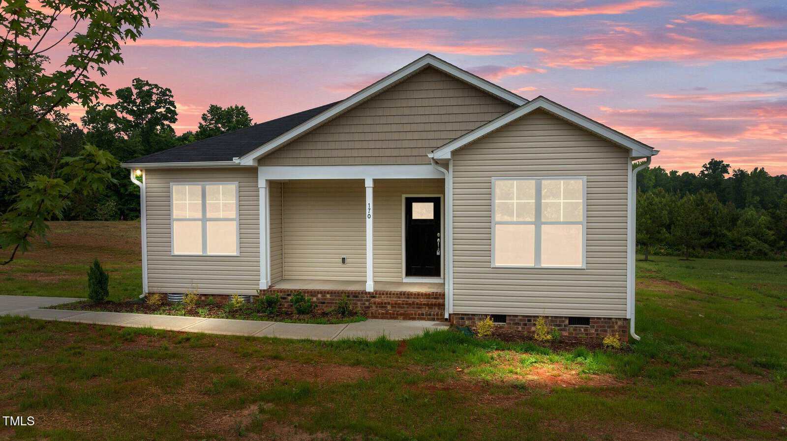 A single-story beige house with white trim and a covered front porch is shown at sunset. The house has a black front door, brick foundation, and sits on a grassy lot with trees in the background.