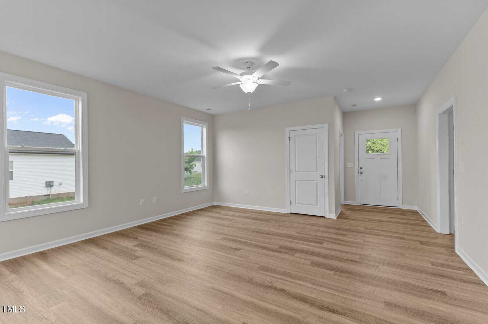Bright, empty room with light wood flooring, beige walls, two windows, a ceiling fan, a white door, and a white front door with a window, all seen in natural daylight.