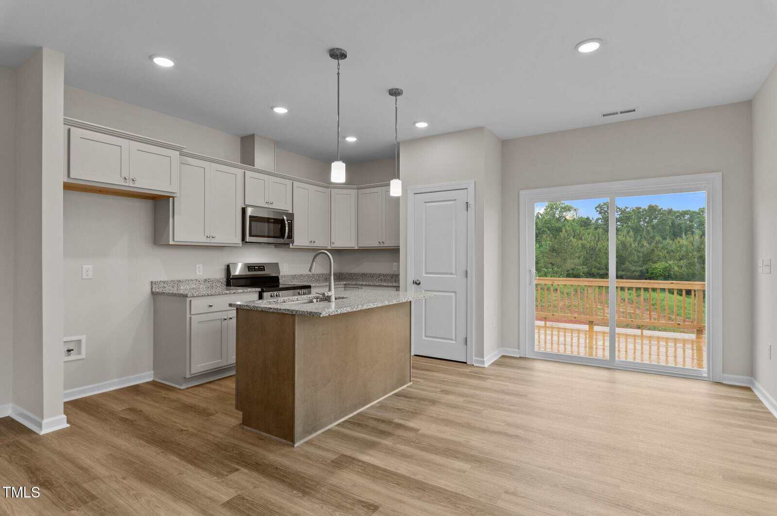 Modern kitchen with light gray cabinets, granite countertops, stainless steel appliances, a center island with a sink, pendant lights, and sliding glass doors leading to a wooden deck with a view of trees.