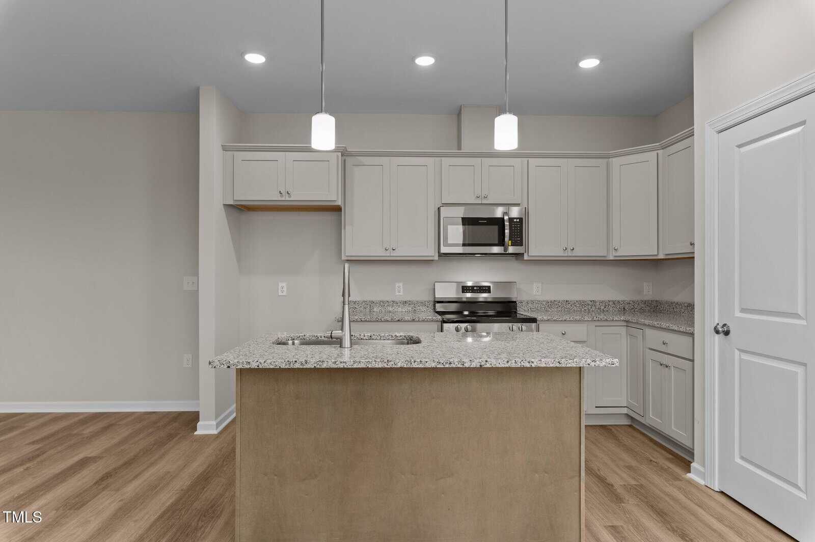 A modern kitchen with white cabinets, granite countertops, a center island with sink, stainless steel appliances including a microwave and stove, pendant lighting, and wood-style flooring.