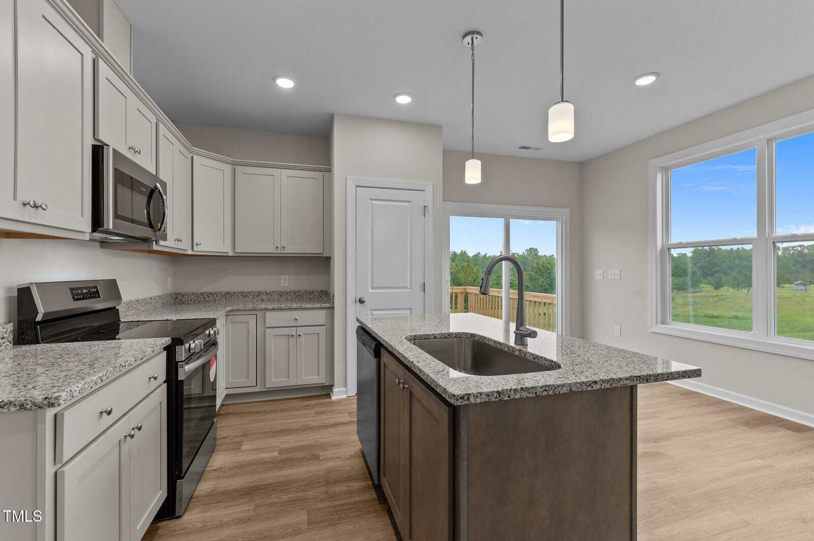 Modern kitchen with stainless steel appliances, granite countertops, an island with a sink, wood flooring, white cabinets, pendant lighting, large windows, and a door leading to a deck with a view of greenery outside.